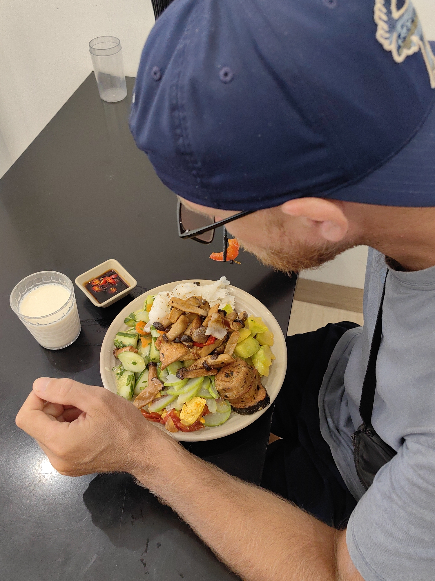 A man wearing a blue baseball cap is seated at a dark table in what appears to be an indoor dining area in Hong Gai Ward, Vietnam. In front of him is a bowl of food containing rice noodles, mushrooms, zucchini, potatoes, and what looks like fried tofu. There is also a small dish of chili sauce and a glass of a white liquid (possibly milk or soy milk) on the table. The man is looking down at the food with what appear to be glasses perched on his nose or mouth. He is wearing a light blue shirt, and a small black bag is slung across his body.