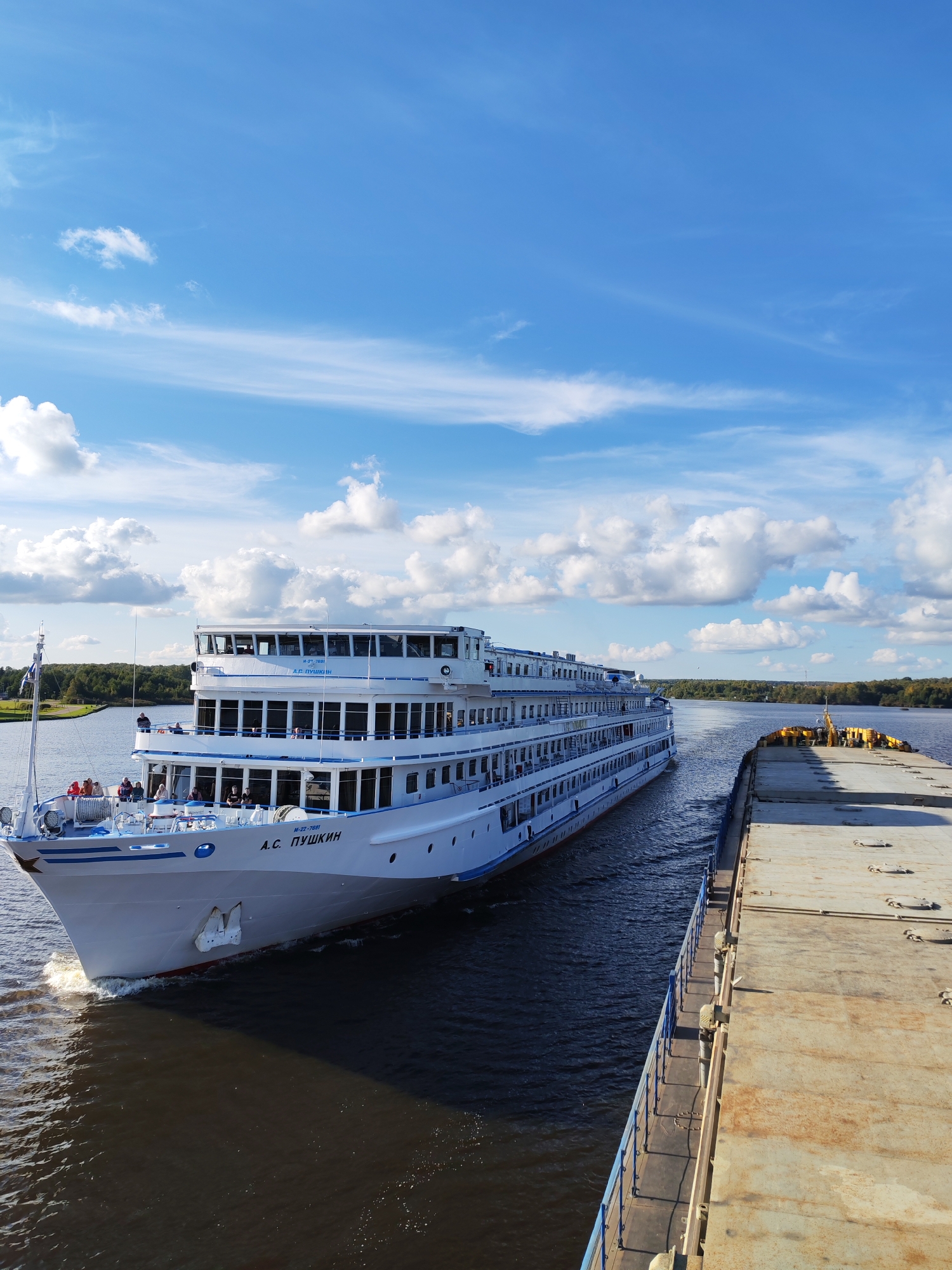 A wide shot captures a white passenger ship, the "A.S. PUSHKIN," sailing on a dark river under a bright blue sky with scattered white clouds. The ship, featuring multiple decks with rows of windows and people visible on its upper decks, is positioned in the left foreground. To the right of the passenger ship, a long, flat-topped barge with a rusty surface and yellow cargo covering is traveling in the same direction. The barge's blue railing separates it from the water. The river flows towards a distant, tree-lined shore under the expansive sky. The scene appears to be a sunny day in the city of Pestovo, Russia, with the river serving as a transportation route for both the passenger vessel and the cargo barge.