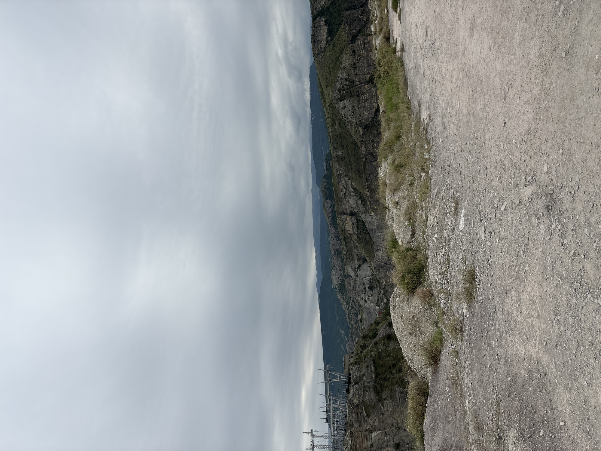 The image depicts a rugged, mountainous landscape with a cloudy sky. In the foreground, there is a dirt path with gravel and sparse vegetation. In the middle ground, the terrain becomes steeper, with rocky outcrops and greenery. To the left, a power substation with tall metal structures and electrical equipment is visible, situated on the edge of a canyon. The background features rolling hills and mountains stretching towards the horizon, under a heavily overcast sky. The scene suggests an industrial area integrated into a natural, remote environment, possibly near the Chirkey Dam in Chirkey, Russia.