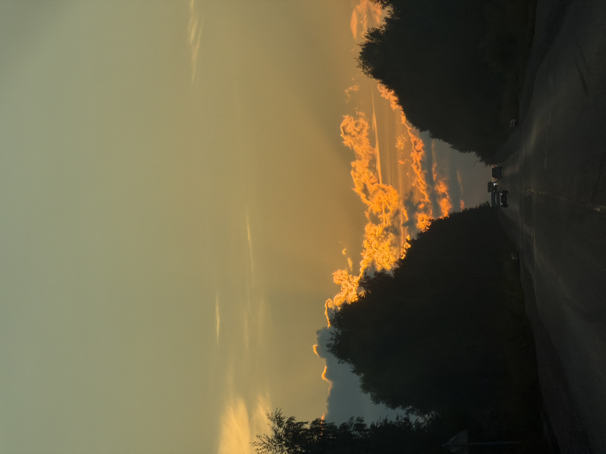 The image depicts a road in Plamya, Russia, at sunset. The sky is filled with hues of orange and yellow, with clouds illuminated by the setting sun. Trees line both sides of the road, casting dark silhouettes against the colorful sky. Several vehicles are visible on the road, traveling in the same direction. The road surface appears to be asphalt, and there is a faint indication of lane markings. A white road sign is partially visible on the left side of the road. The overall impression is of a serene, late-day drive through a rural or semi-rural landscape.