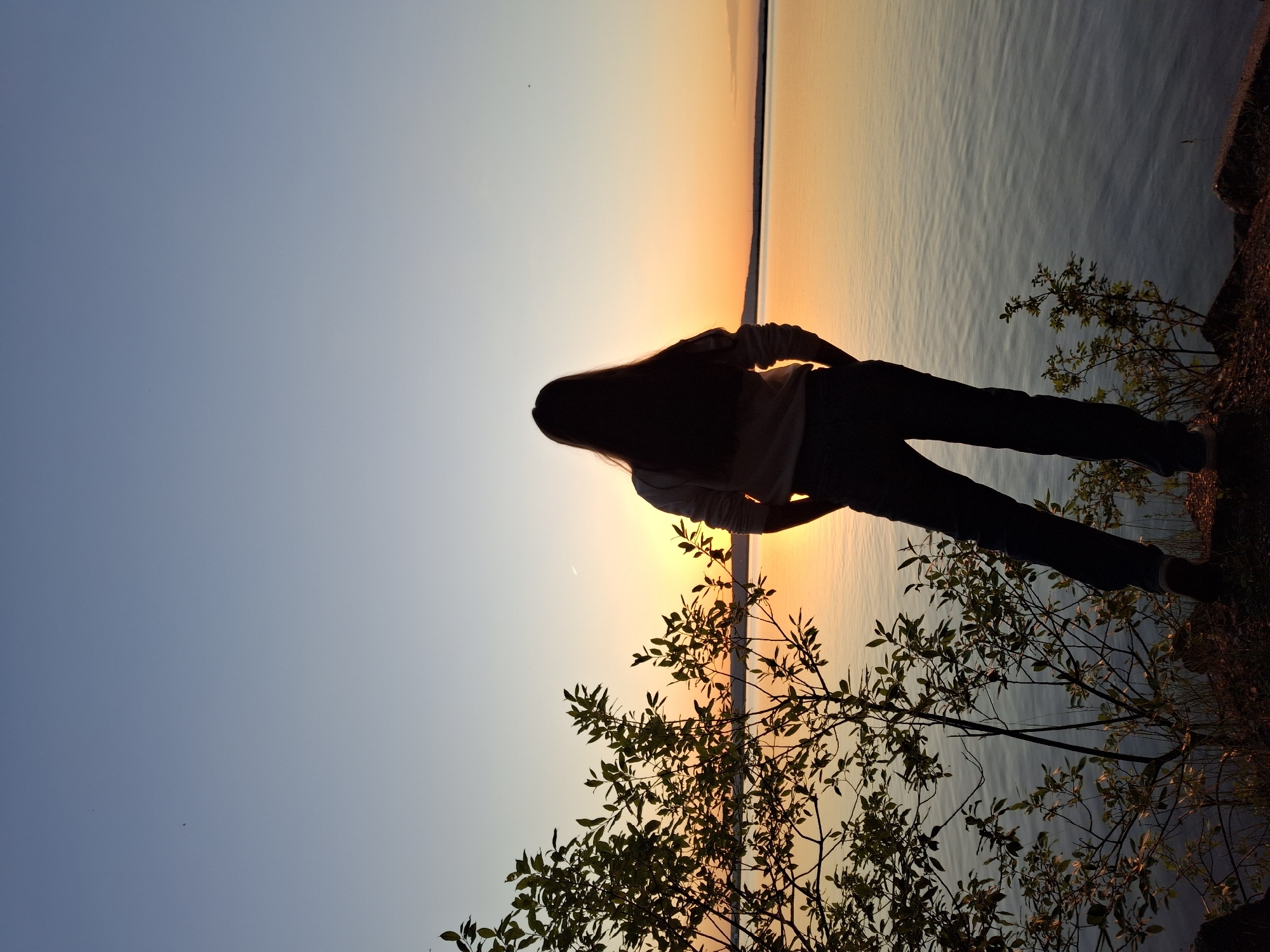 A full shot captures a person standing with their back to the camera, observing a sunset over a calm body of water. The person, silhouetted against the vibrant orange and yellow hues of the setting sun, has long, dark hair and is wearing a light-colored sweatshirt and dark pants. The individual's hands are tucked into their pockets. To the left of the frame, a leafy bush with thin branches is silhouetted against the sunset. The water's surface is smooth, reflecting the colors of the sky. The horizon is visible in the distance, with land faintly appearing beyond the water. The sky transitions from a deep blue at the top to lighter shades of blue and yellow closer to the horizon. The overall scene depicts a serene moment by the water during sunset, likely at the Kama River embankment in Chaikovsky, Russia, given the context of the location.