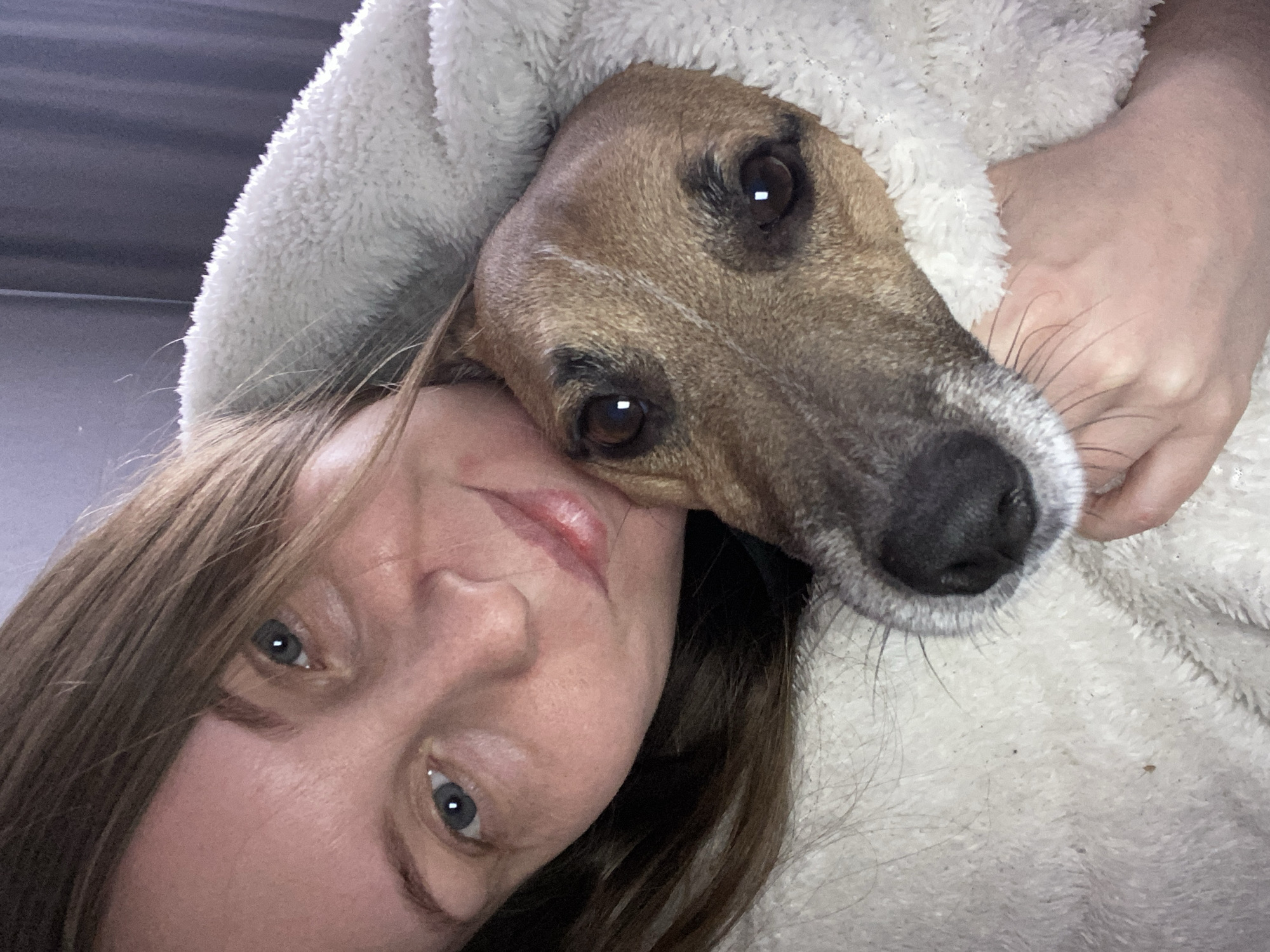 A close-up selfie shows a woman and a dog. The woman has light skin, light brown hair, and blue eyes. Her face is tilted to the left. The dog, a brown and white breed, is positioned to her right, snuggled against her. Both the woman and dog appear to be nestled inside a white, fluffy garment, possibly a robe or blanket. The environment is indistinct, with only a suggestion of a grey surface in the background. The location of Noviy, Russia is not evident within the frame.