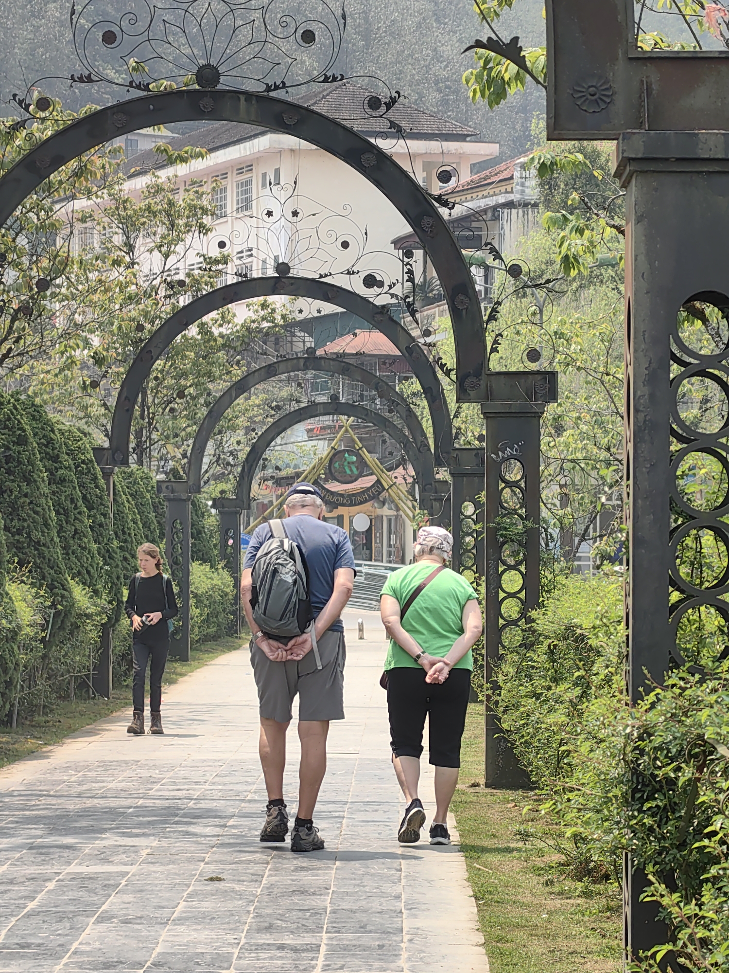 The image depicts an outdoor pathway in Sa Pa, Vietnam, characterized by a series of dark metal archways with decorative scrollwork. Three individuals are present on the paved pathway under clear daylight conditions.
In the foreground, an adult male and an adult female walk away from the camera. The male, positioned left of center, wears a blue t-shirt, gray shorts, a blue baseball cap, gray hiking boots, and a gray backpack. His hands are clasped behind his back. To his right, the female wears a green t-shirt, black capri pants, dark sneakers, and a light-colored head covering; her hands are also clasped behind her back, with a maroon strap visible across her back.
Further down the pathway on the left side, a young adult female walks towards the camera. She wears a black long-sleeved shirt, black pants, and brown hiking boots, holding a small black object in her hands.
The pathway is composed of rectangular gray paving stones and is flanked by tall, neatly trimmed green hedges on the left and shorter green bushes and grass on the right. Numerous ornate dark metal archways, featuring floral motifs and circular cutouts on their supporting columns, line the path, receding into the distance. Beyond the archways, multiple multi-story buildings with various architectural styles are visible, including a prominent light-colored structure with numerous windows. A sign with Vietnamese text, likely reading "ĐƯỜNG TINH YÊU" (Love Road), and a triangular emblem is discernible further down the path. Green trees and foliage are interspersed throughout the scene, with a hazy, mountainous background suggesting the topography of Sa Pa.