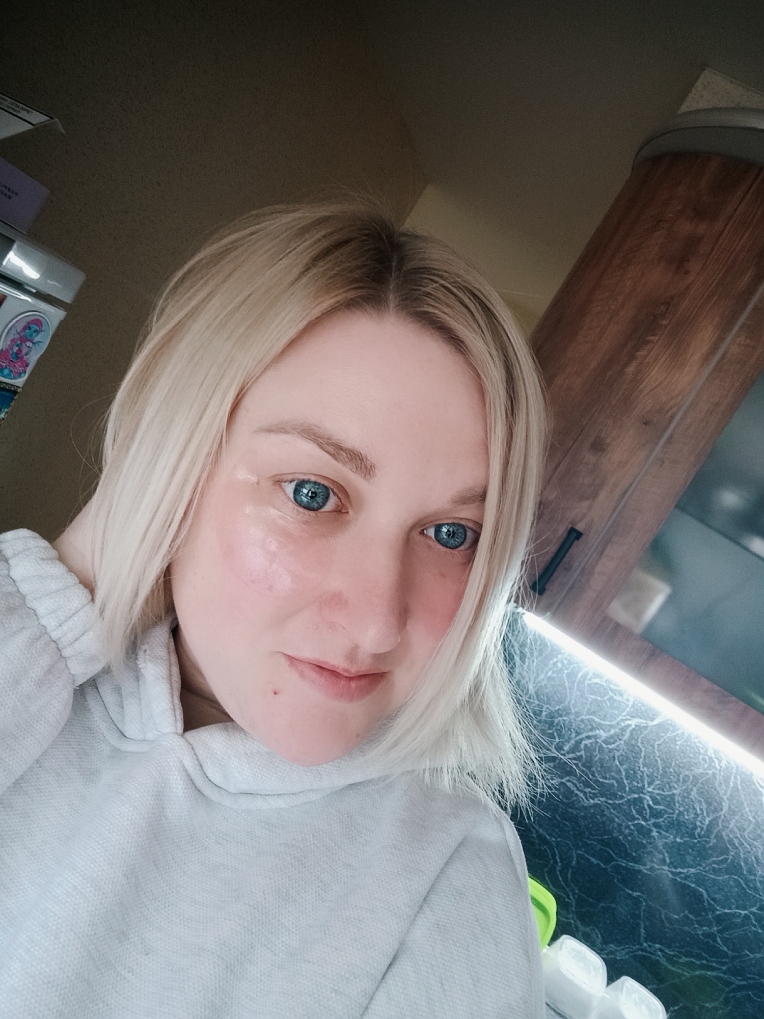 A young woman with blonde hair and striking blue eyes is depicted in a close-up, upward-angled selfie, likely taken indoors in Chaikovsky, Russia. She has a serene expression with a subtle smile and appears to be wearing under-eye patches, suggesting a skincare routine or relaxation. She is dressed in a light grey hooded sweatshirt.
The background reveals parts of what appears to be a kitchen or a room with a kitchenette. To her left, a white container with a colorful, possibly cartoonish, blue and pink sticker is partially visible, along with a glimpse of a purple box above it, on which "artes" and some illegible text can be faintly seen. To her right and behind her, dark wooden cabinetry is visible. Below her chin, a dark, textured countertop, possibly a stone or patterned laminate, is illuminated by a bright, linear strip light mounted underneath the cabinet. Some small, clear, possibly plastic containers or ice cube trays are also on the countertop. The lighting suggests an indoor setting, possibly during the day or evening. The overall mood is calm and personal.