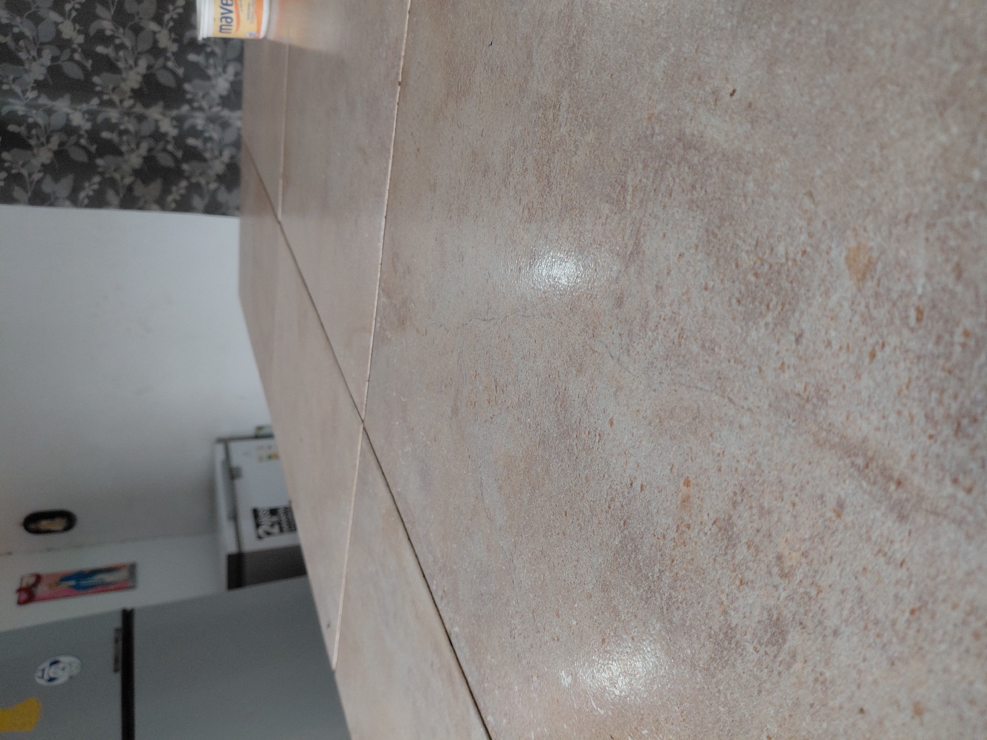 The image displays an indoor scene, likely a kitchen or utility area, with a tiled surface dominating the foreground. The tiles are light beige or tan, featuring irregular brown and orange speckles, and appear to have a slightly textured, matte finish with some glossy reflections. Distinct grout lines indicate individual tiles.
In the background, on the left side, a wall is covered with dark grey or black wallpaper featuring a pattern of leaves or foliage on a lighter grey ground. Above this wallpaper, a white container with orange lettering is partially visible; the text appears to read "maves" or "maven." Below the wallpaper, a plain, light-colored wall section is visible.
To the right in the background, a residential interior space is seen. A grey refrigerator with visible magnets or stickers occupies the lower portion of this area. Above it, on the wall, there is a small, colorful, rectangular picture or decoration. Further back, another white appliance, possibly a second refrigerator or a cabinet, is visible, marked with dark labels or graphics. An oval-shaped light fixture is mounted on the white ceiling above.
No specific activity is taking place, and there are no people in the frame. The lighting suggests an indoor setting, possibly during the day due to the ambient light, but specific time or weather cannot be determined. The scene offers a glimpse into a domestic space in Santa Ana, Venezuela.