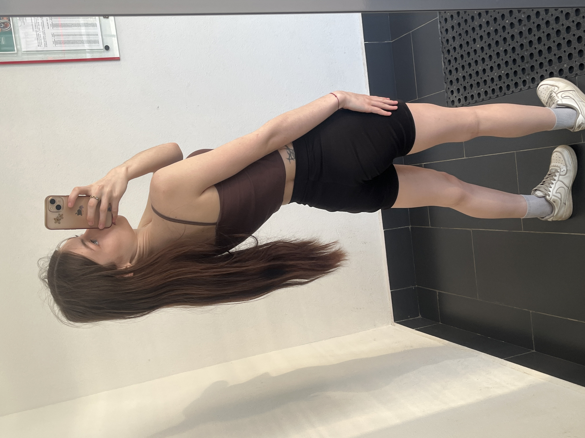 A young individual with long, straight, dark brown hair is captured in a full-body mirror selfie, standing in an indoor setting. The person is wearing a dark brown spaghetti strap crop top, black shorts, white athletic shoes, and light grey socks. A light-colored smartphone with a floral pattern case is held up by the right hand to take the photo, with a silver ring visible on the left ring finger and a red string bracelet on the right wrist. A tattoo is present on the left ribcage area. The background features plain white walls and a black tiled floor with white grout. A black tile baseboard runs along the bottom of the wall. On the right side of the mirror's reflection (left in the room), a glass-covered bulletin board containing several documents, some with red lines, is mounted on the wall. A textured black rubber mat is visible on the floor to the right side of the image. The specific location in the city of Unknown, Russia, is not identifiable from the visual content.