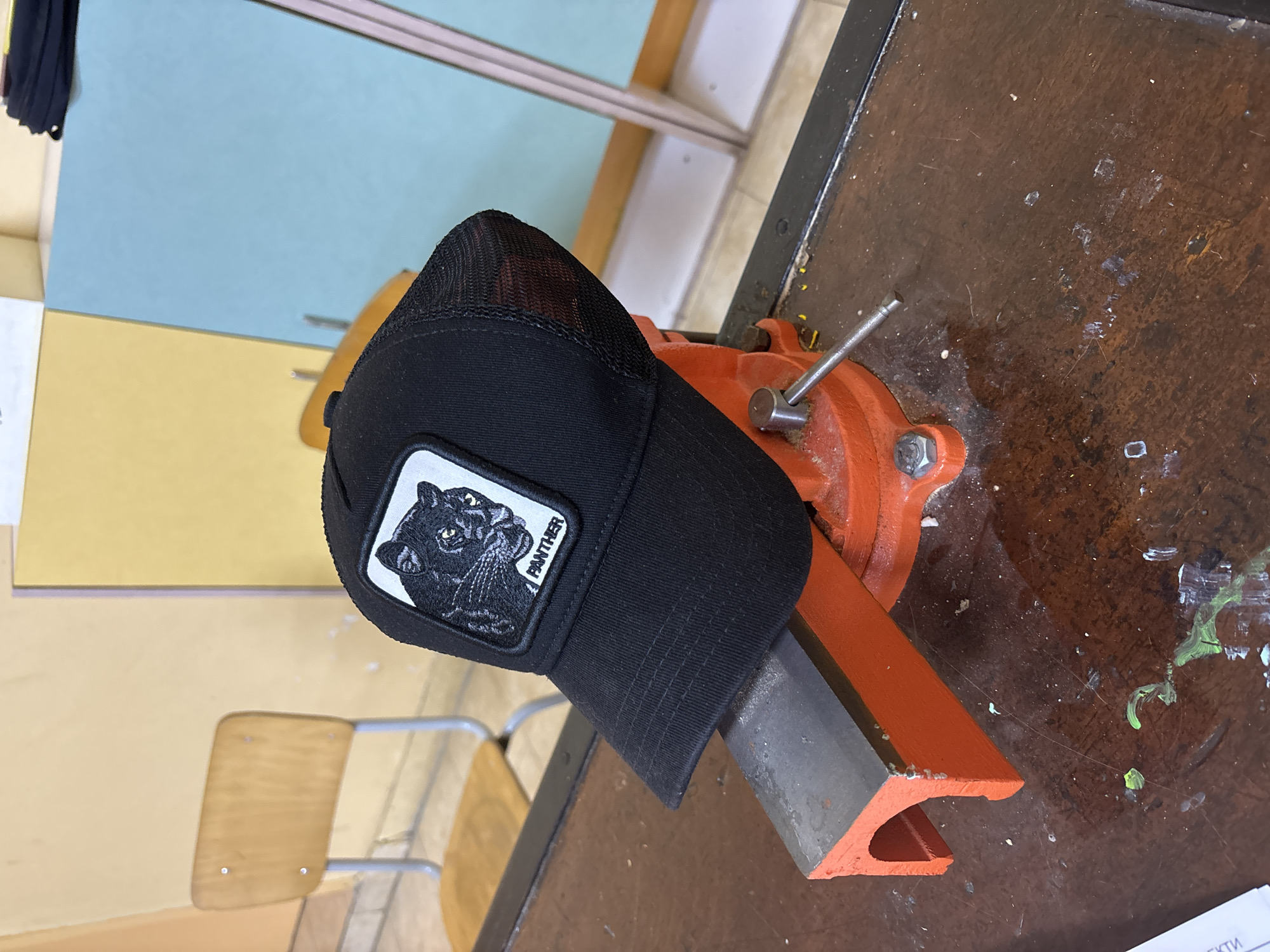 This image, captured in Ruse, Bulgaria, features a black trucker-style baseball cap prominently displayed, resting on the jaws of an orange and grey bench vise. The cap is made of black fabric, with a mesh back and a solid front panel. A white square patch is sewn onto the front, depicting a detailed black panther's head. Below the panther's image, the word "PANTHER" is written vertically in small, black capital letters.
The orange metal vise is clamped onto a dark, worn wooden workbench or table. The surface of the table is scratched, dusted, and marked with splatters of light green and white paint, suggesting a workspace. The hat is placed over a grey, possibly metal, block held by the vise jaws.
In the background, a light blue wall rises from a lighter yellow-beige wall or surface. Part of a light-colored wooden chair back with a silver metal frame is visible on the left. A horizontal metal bar or rail crosses the upper background. The setting suggests an indoor workshop, classroom, or practical area. There is no discernible activity taking place, nor any specific time of day or emotions evident. A fragment of paper in the bottom right corner shows some partially legible Cyrillic text, with the letters "ЕКТИ" visible.