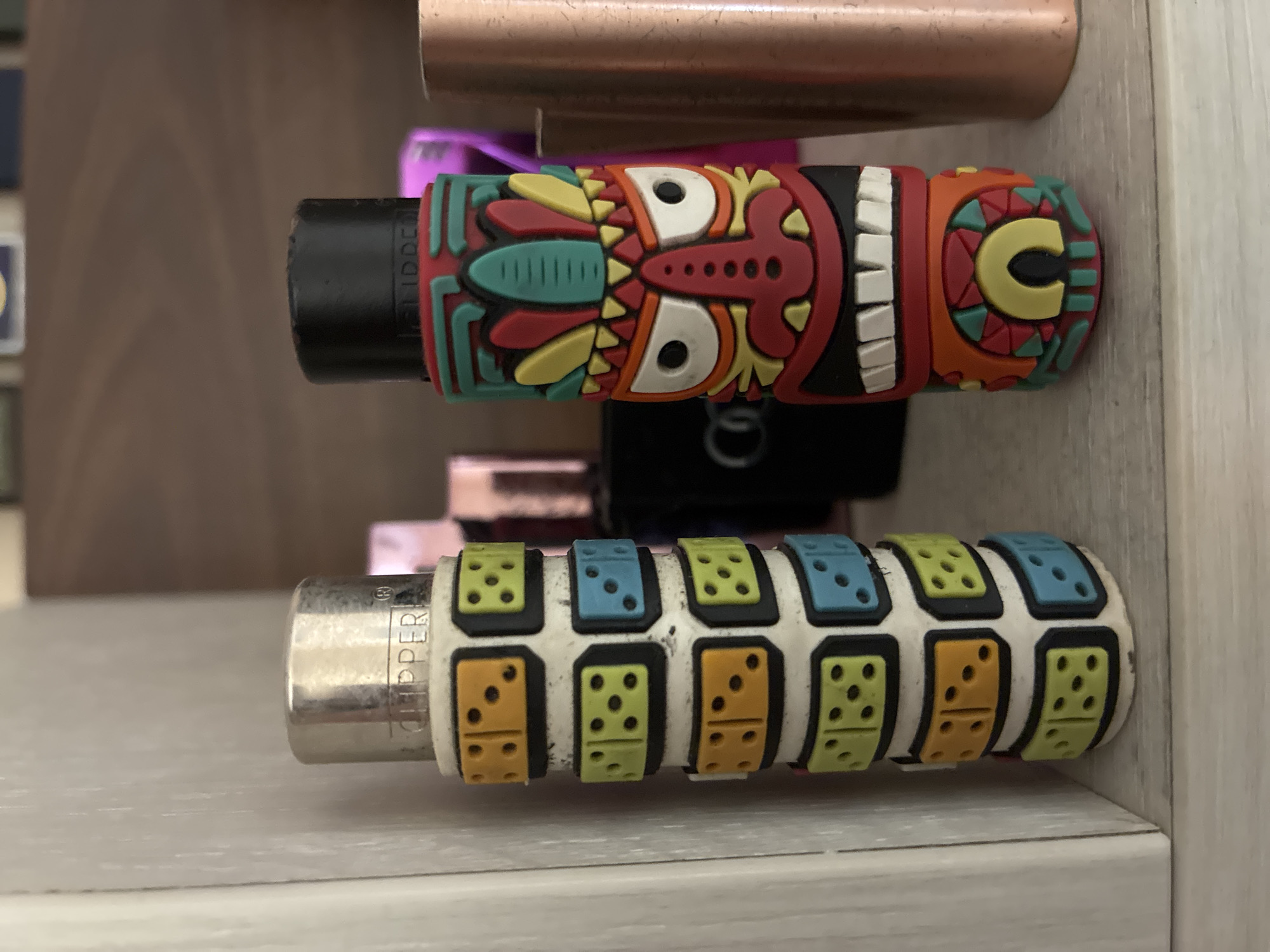Two cylindrical objects, likely lighters or similar handheld devices, are displayed horizontally on a light-colored surface, possibly a shelf or cabinet. A blurred, dark wooden surface serves as the background, with hints of other objects in the far distance.
The top object features a colorful, rubbery sleeve resembling a stylized tiki mask or an Aztec-inspired face. This design incorporates vibrant shades of red, yellow, black, white, turquoise, and orange, depicting large eyes, a prominent nose, and a wide mouth with teeth. The patterns are intricate and symmetrical. On the black, exposed end of this object, the word "CLIPPER" is visible in white lettering.
The bottom object has a white, rubbery sleeve adorned with rectangular domino-like patterns arranged in a grid. These domino pieces come in distinct colors: yellow, light blue, green, and orange, each featuring black dots to represent numbers (e.g., 2, 3, 5, 6). The metallic, worn end of this object clearly shows the word "CLIPPER" engraved, along with a registered trademark symbol (®) and possibly another small mark.
A metallic, copper-toned cylinder is partially visible at the very top of the frame, resting vertically. The overall scene suggests an indoor setting, possibly a personal collection or storage area. No activity is depicted, and there are no discernible cues for time of day, weather, or emotions.