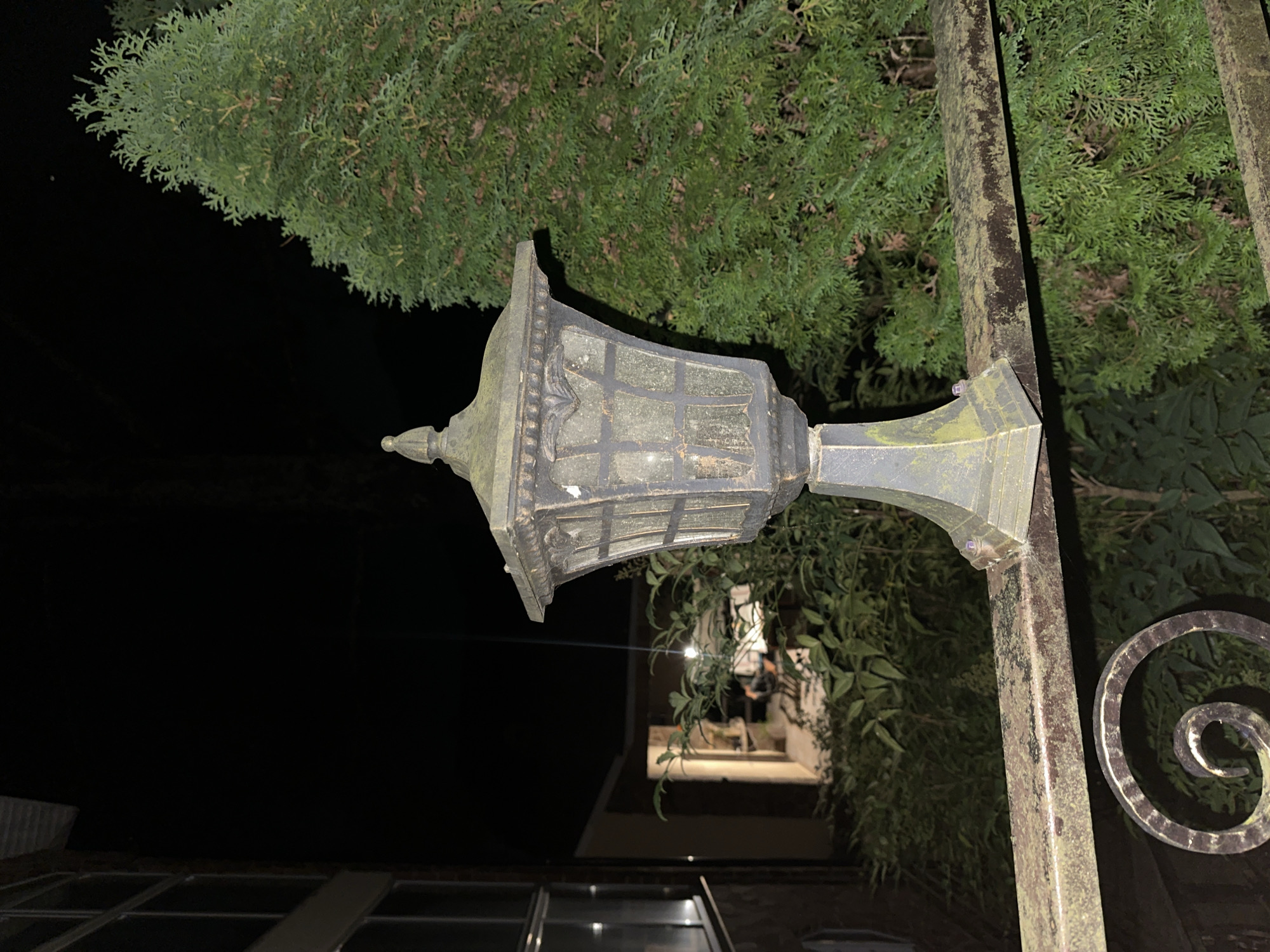 An ornate, dark-colored outdoor lantern with a hexagonal glass paneled body and decorative finial stands on a weathered, moss-covered metal railing at night. The railing is part of a fence or balcony. In the background, a large evergreen tree with dense foliage is visible to the right, while a building with windows is partially visible on the left. There are no people or animals in the frame. The scene is illuminated by an unseen light source, possibly the lantern itself. The overall impression is one of an exterior nighttime setting, likely in a residential or public outdoor area. No specific features definitively place this image in Sochi, Russia.