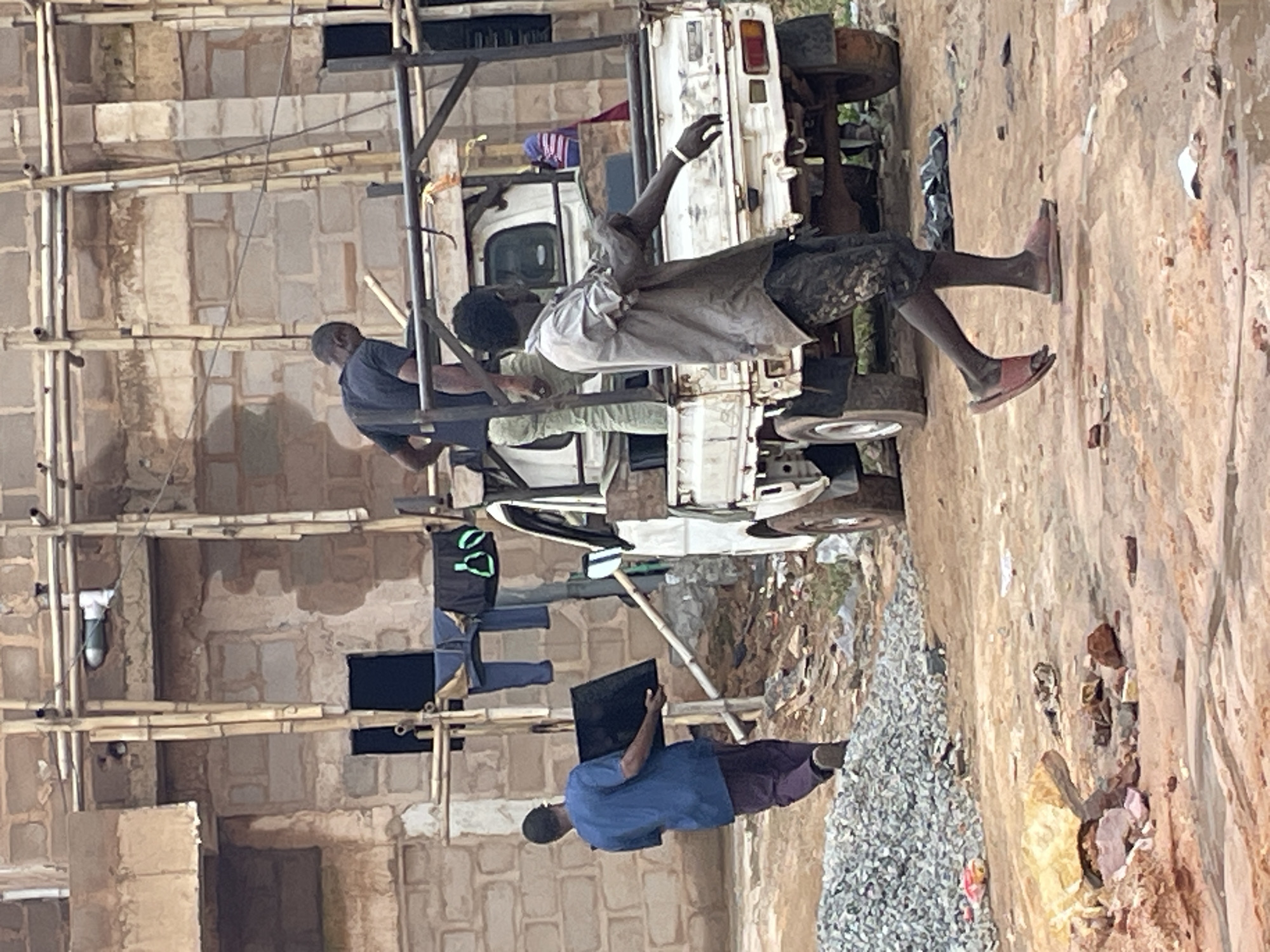 The image depicts a construction site in Enugu, Nigeria. A man in a blue shirt and dark trousers is carrying a black flat object, possibly a television or screen, towards a white pickup truck. Another man in a grey shirt and patterned shorts is standing near the back of the truck. A third man in a dark t-shirt and light-colored pants is visible in the background, standing on scaffolding. Clothes are hanging on a makeshift clothesline behind the men. The environment is dusty and appears to be undergoing construction, with piles of gravel and debris visible in the foreground.