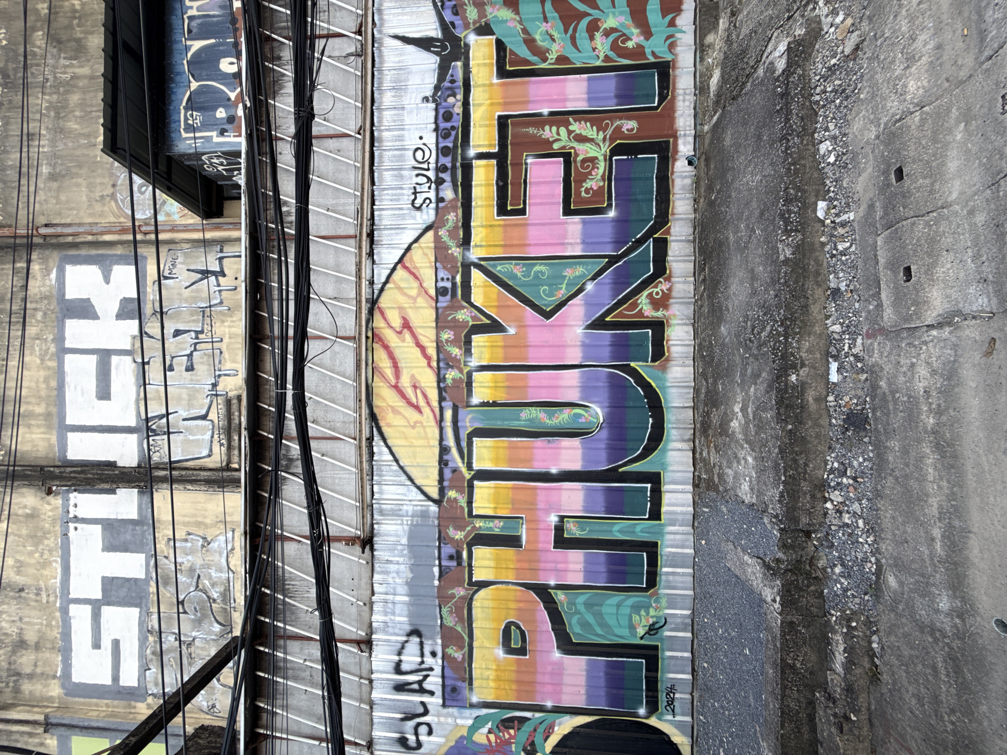 The image shows a close-up of a graffiti mural on a corrugated metal wall in Phuket City, Thailand. The mural features the word "PHUKET" rendered in large, colorful, block-style letters with a gradient of purple, pink, yellow, and orange. The letters are outlined in black and have decorative elements of green vines and pink flowers. Above the main mural, there are other graffiti tags and drawings, including the word "STICK" in large white letters and other scribbled tags. A section of a dark blue and black graffiti tag is visible on the right. Electrical wires are strung across the upper part of the mural. The bottom of the image shows a rough, concrete sidewalk and a drainage grate. The overall scene depicts an urban environment with significant graffiti art.