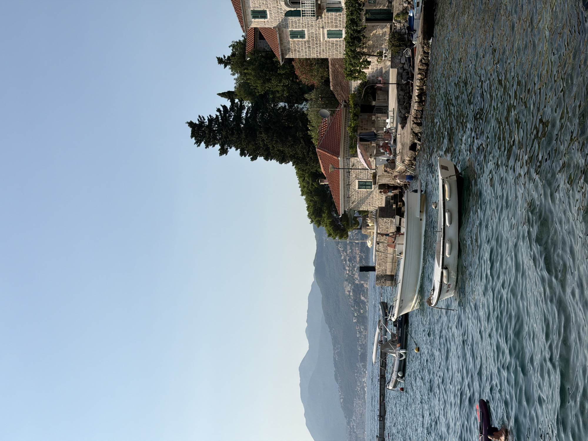 This is a vertical, eye-level, medium shot of a waterfront scene. The left side of the image features a clear, light blue sky, with the bottom portion of a tall, dark green coniferous tree visible. To the right of the sky and tree, a stone building with a red tiled roof is situated along a waterfront. The building has multiple windows with green shutters and a stone facade. In front of the building, there are several outdoor seating areas with chairs and tables, and a few people are visible, appearing to be relaxed.
Adjacent to the building is the water, which is a deep blue with noticeable ripples and reflections. Several boats are moored along the waterfront. A long, white boat with a cabin is the most prominent. Two smaller, inflatable boats are tied to a darker boat. A person is partially visible in the water on the far right, with a purple object nearby.
In the background, across the water, are layered mountains covered in greenery, with buildings scattered on their slopes, suggesting a coastal town or village. The lighting suggests it is daytime, likely late afternoon or early evening, given the warm light and clear sky, with a gentle breeze causing the ripples on the water. The scene evokes a sense of tranquility and a Mediterranean coastal atmosphere. Some faint text is visible on the side of a boat, but it is too blurry to read. The location context provided is Zelenika, Montenegro.