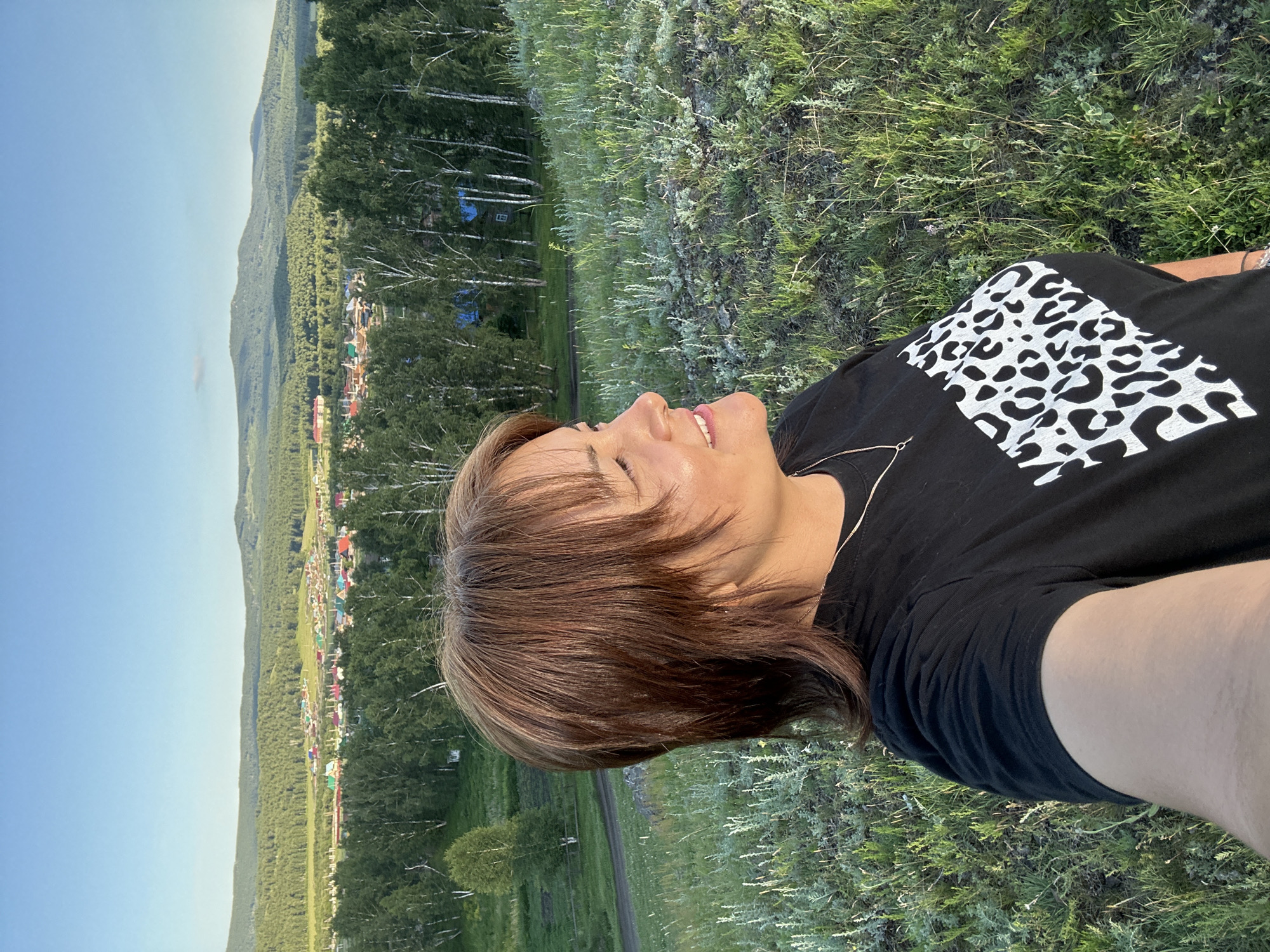 A woman with shoulder-length brown hair is taking a selfie outdoors on a grassy hillside. She is wearing a black t-shirt with a white leopard print design. She is looking to her right, with a slight smile. In the background, there is a village nestled in a valley, surrounded by a lush green forest and rolling hills under a clear blue sky. The sunlight suggests it is late afternoon or early evening.