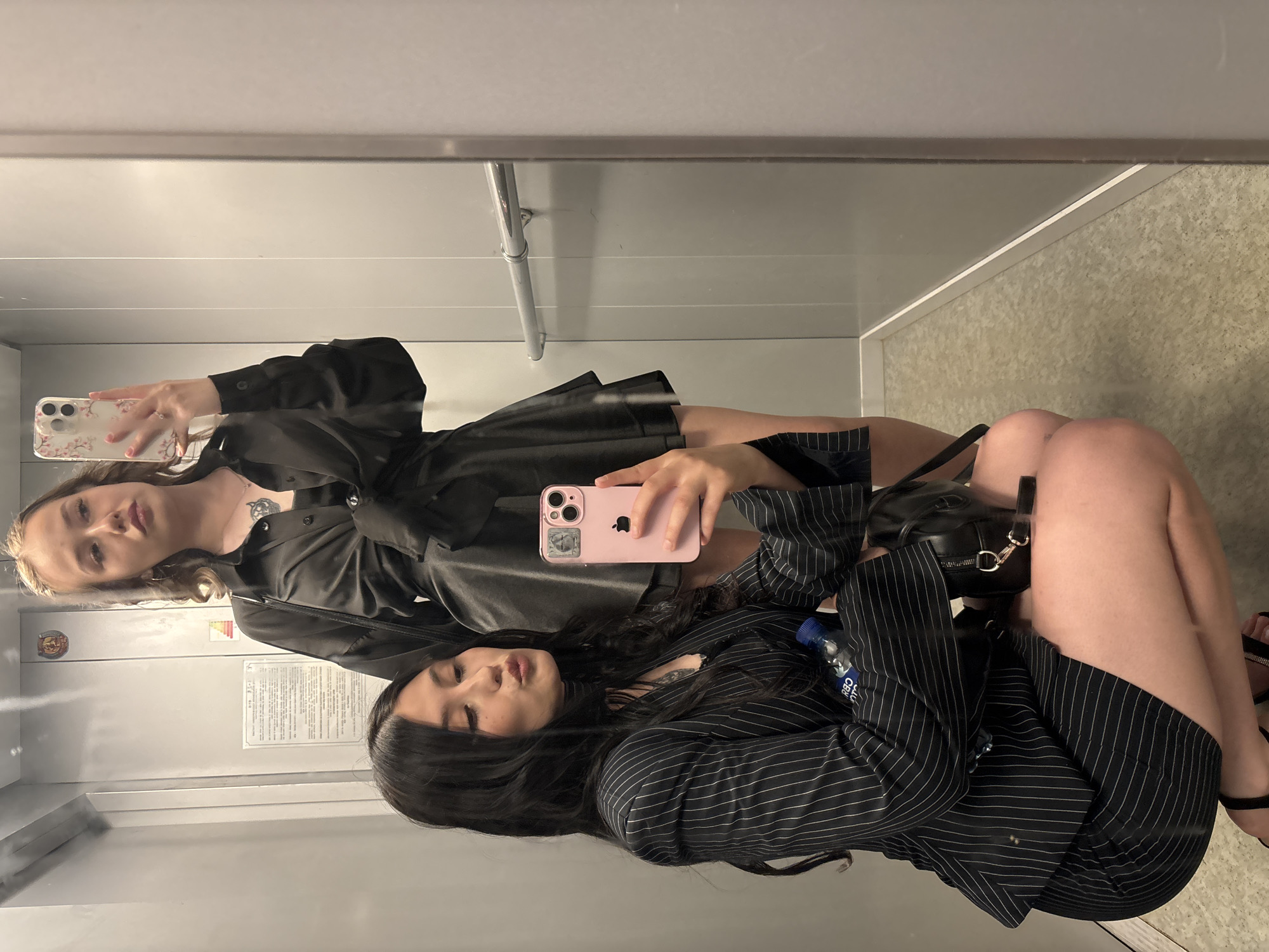 Two young women are posing for a selfie in an elevator. The woman in the foreground, on the left, is crouching and holding a pink smartphone in her right hand, taking the photo. She has long dark hair, is wearing a black pinstripe blazer over a black top and a black skirt. A blue water bottle and a small black bag are visible near her. The woman in the background is standing and holding a phone in her right hand, also taking a selfie. She has blonde hair and is wearing a black satin-like shirt tied at the waist and a black skirt. The elevator walls are grey and metallic. There is a handrail on the right wall. The floor of the elevator is a speckled grey.