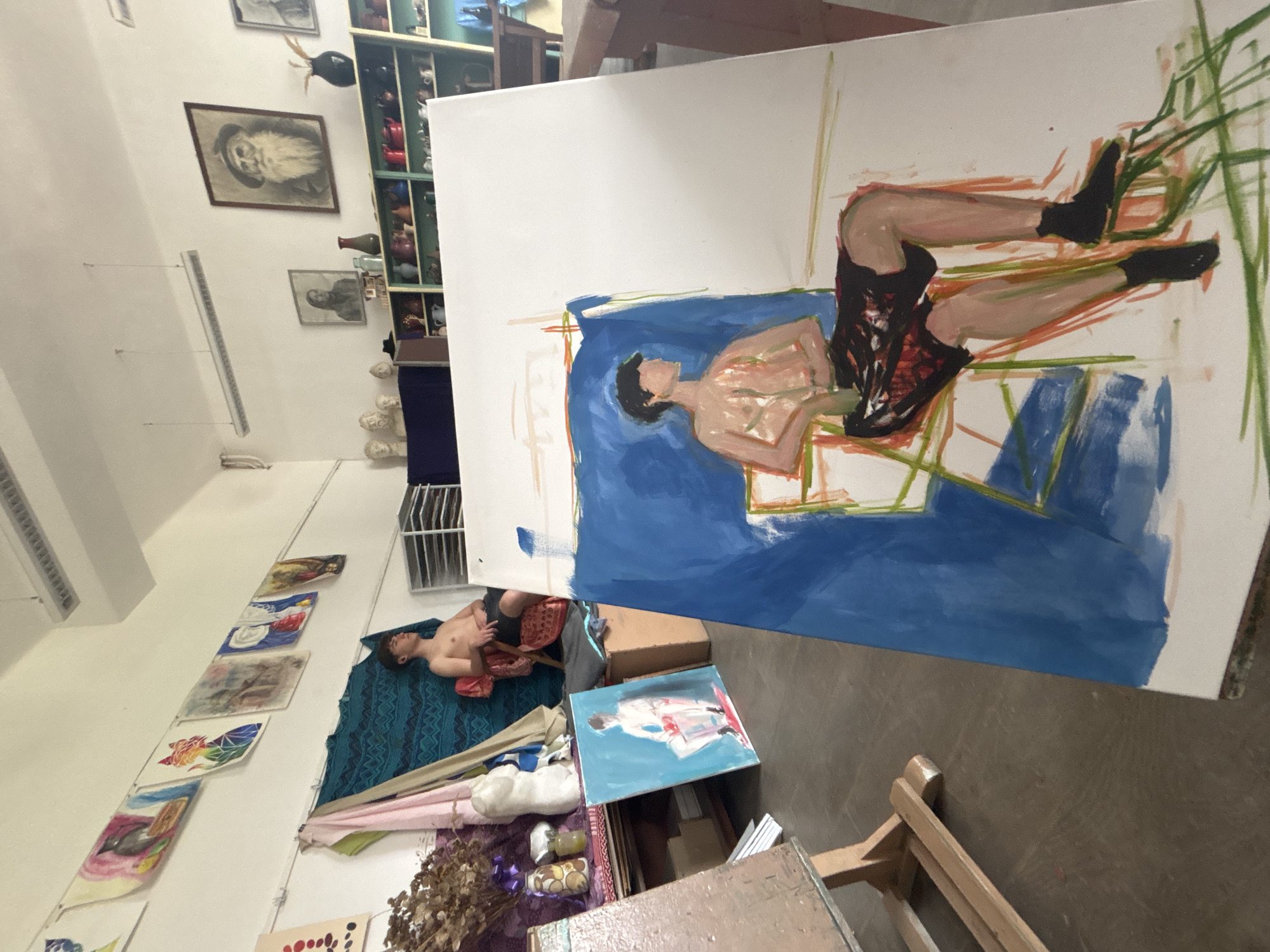 The image shows an artist's studio. A partially finished painting on a canvas depicting a reclining figure in shorts and black socks is the foreground focus. The canvas sits on an easel, whose legs are visible. In the background, on a patterned blue blanket, is a person reclining, presumably the model for the painting. The room has other paintings displayed on the wall. Various art supplies and objects like boxes, fabric, dried flowers, and an unfinished painting are scattered around the lower part of the frame. A bookshelf filled with items and framed images are visible on the upper left. Based on the information provided, the location is in the city of Troyan, Bulgaria.
