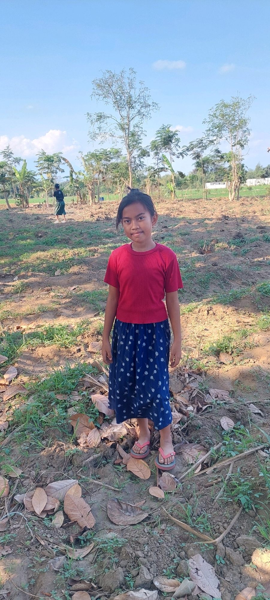 A young girl with dark hair pulled back into a ponytail stands facing the camera in an outdoor, rural setting in Rogotrunan, Indonesia. She is wearing a red short-sleeved shirt and a dark blue skirt with a white polka dot pattern. She has on pink flip-flops. The ground is dry and consists of dirt, scattered leaves, and some sparse green vegetation. In the background, there are several trees, some with sparse foliage, and other vegetation. A person in dark clothing is walking away from the camera in the mid-ground to the left. The sky is clear blue with a few wispy white clouds.