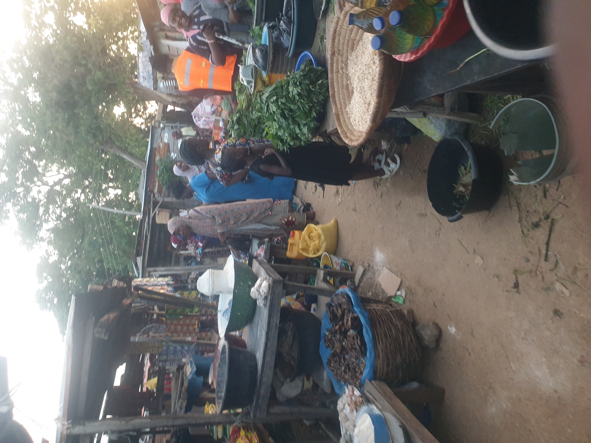 The image depicts an outdoor market scene in an unidentified location. Several individuals are present, engaged in buying and selling activities. There are various goods on display, including fresh produce such as leafy greens and dried goods stored in baskets and buckets. The environment appears to be a dirt pathway surrounded by makeshift stalls or tables constructed from wood and metal. The lighting suggests it is daytime, with strong sunlight casting shadows.
