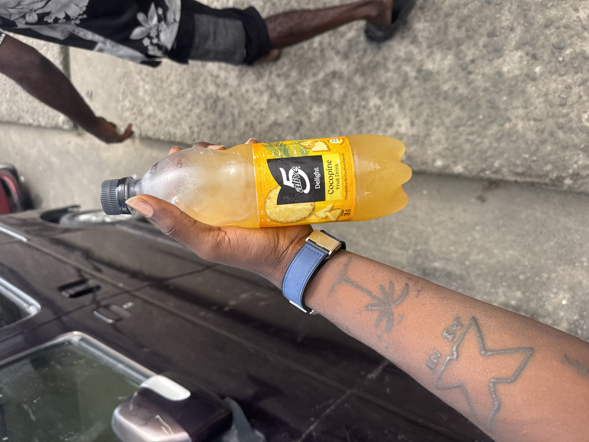 A person is holding a bottle of 5 Alive Delight Cocopine Fruit Drink, pineapple with coconut flavor, outdoors. The person is Black and has tattoos on their arm, including a palm tree and a star, and wears a blue watch. Part of a black car is visible in the foreground. The person appears to be standing on a sidewalk in Port-Harcourt, Nigeria. Other parts of a person are visible in the background.