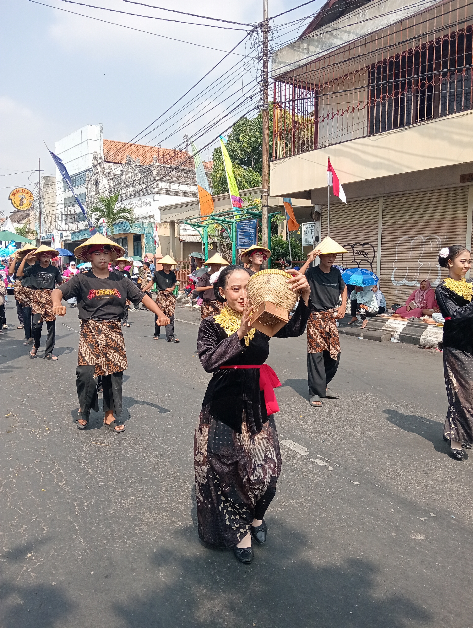 This is a full-frame, eye-level, outdoor photograph of a cultural parade in the city of Demak, Indonesia, on a bright, sunny day. The parade participants are walking on an asphalt street lined with buildings and overhead electrical wires.
In the foreground, a young woman dressed in a black velvet outfit with gold trim and a red sash is performing a traditional dance. She is holding a woven basket filled with objects. To her right, another young woman in a similar outfit with gold trim and a white flower in her hair is also dancing.
Behind them, a group of young men are marching in unison, also dressed in traditional attire. They wear conical straw hats, black t-shirts, and patterned sarongs over black pants. The man in the center is holding his hat, looking towards the viewer. Another man to his left is holding his hat as well.
In the background, more parade participants can be seen, along with spectators sitting on the side of the street, some under umbrellas. Buildings with traditional Indonesian architecture, some with ornate details and barred windows, line the street. Flags, including the Indonesian flag, are displayed along the parade route. Electrical wires are visible crisscrossing the sky. The overall scene depicts a vibrant cultural event taking place in a bustling urban environment.