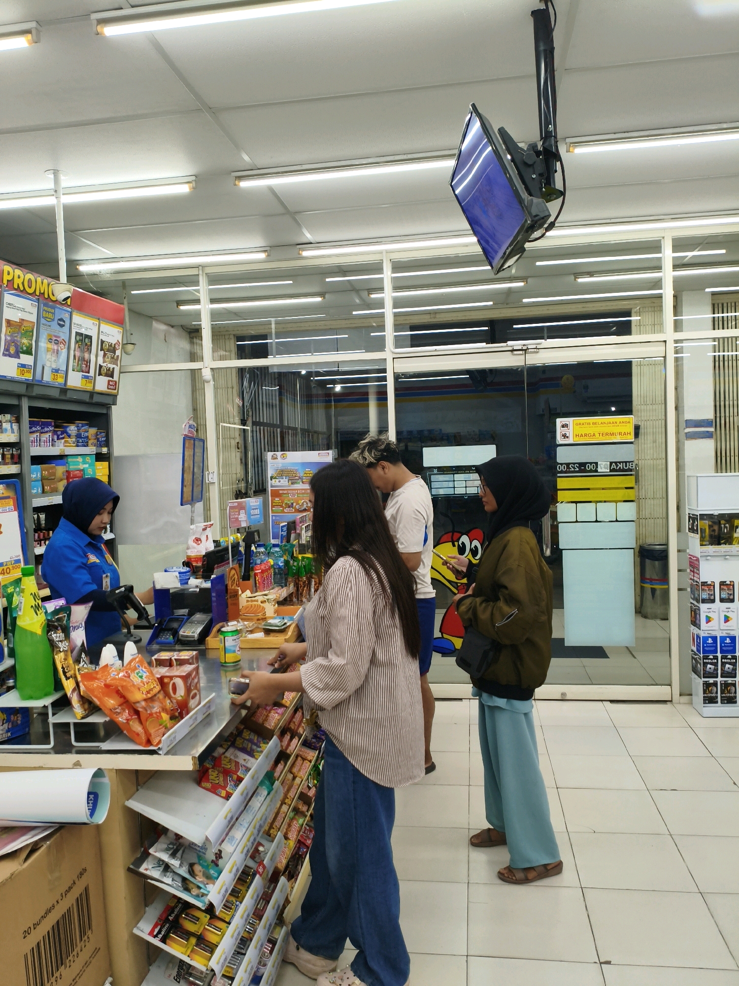 In a convenience store in Sumberejo Wetan, Indonesia, a cashier in a blue uniform with a dark hijab is serving customers at a checkout counter. Two women and one man are visible at the counter. One woman, wearing a striped shirt and blue jeans, is standing on the right, looking down at the counter. Another woman, wearing a dark hijab and a green jacket, is standing to her right, holding a phone and looking towards the left. The man, wearing a white t-shirt, is partially visible behind the woman in the striped shirt. The counter is filled with various products, including snacks, drinks, and other convenience items. Shelves stocked with merchandise are visible behind the cashier. The store appears well-lit, with fluorescent lights on the ceiling. A security camera is mounted on the ceiling, and a monitor is hanging from it. The floor is tiled. A glass door leads to the outside, where more shelves of products are visible.