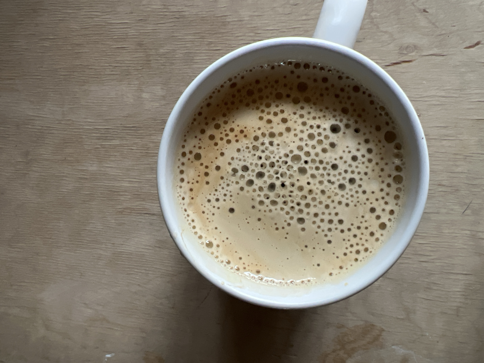 An overhead shot shows a white mug filled with coffee, topped with a thick layer of light brown froth, resting on a light-colored wooden surface. The coffee has numerous bubbles of varying sizes across its foamy surface, indicating it's likely an espresso, cappuccino, or latte. The liquid coffee visible beneath the froth is a darker brown.
The mug is plain white ceramic, and its handle is partially visible in the upper right quadrant of the image. The wooden surface, which takes up the majority of the background, appears to be a table or countertop. It has a visible horizontal grain pattern and a muted, natural tone. There are some faint marks or subtle imperfections on the wood surface, such as slight smudges or light discolorations, particularly noticeable in the bottom left and right areas of the wider image. No people are visible, nor is any specific activity taking place other than the presence of the coffee cup. The time of day, weather, and precise location cannot be determined from the image. There is no visible text.