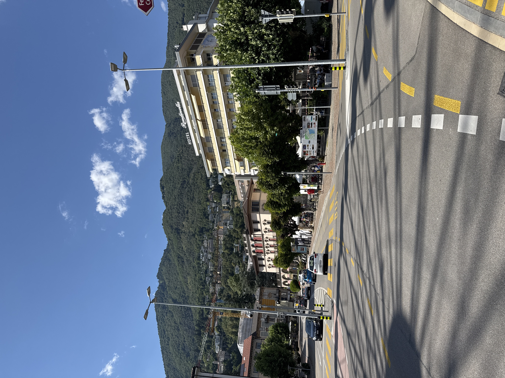 The image depicts a street intersection in Locarno, Switzerland, with a prominent yellow and white striped crosswalk. A multi-story building labeled "HOTEL Rondinella" is visible on the right, nestled against a backdrop of a densely wooded, mountainous hillside. Several cars are driving on the road, and a few pedestrians are present on the sidewalks. Streetlights and traffic signals are also visible. The sky is clear blue with scattered white clouds. The overall scene suggests a bright, sunny day in a European city.