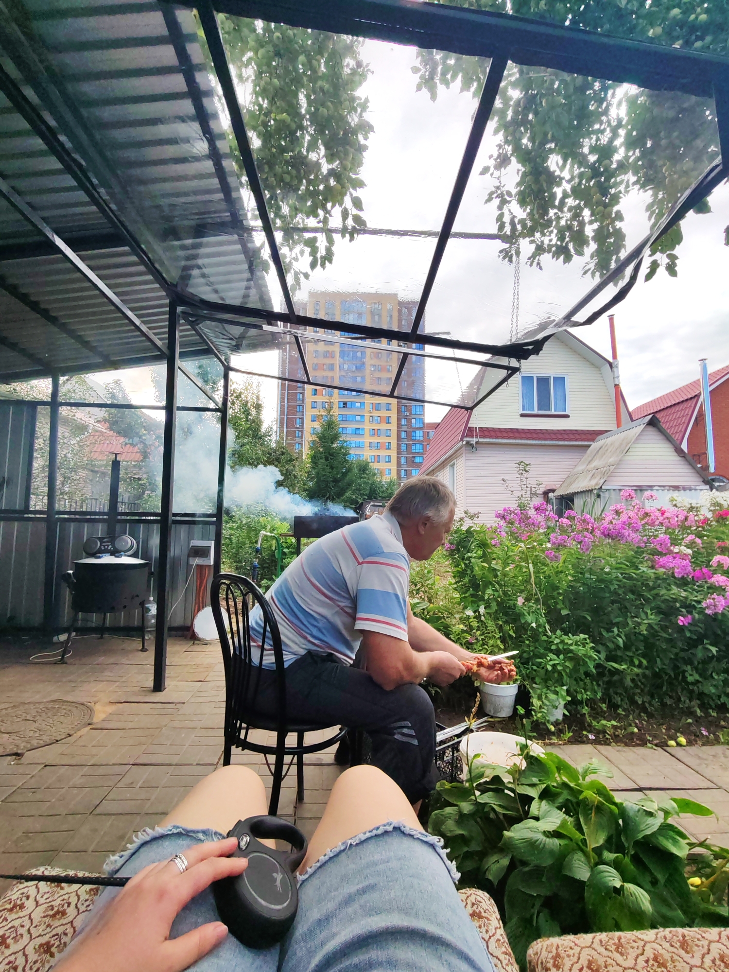 In Pervomaysky, Russia, a man is sitting on a chair under a transparent roof, preparing food on skewers. He is wearing a striped shirt and dark pants. In the background, a large building with multiple floors stands tall. To the right of the man, pink flowers are in bloom. A woman is in the foreground, showing her legs, wearing ripped jeans, and holding a black dog leash. The environment appears to be a backyard with a tiled floor and lush greenery.