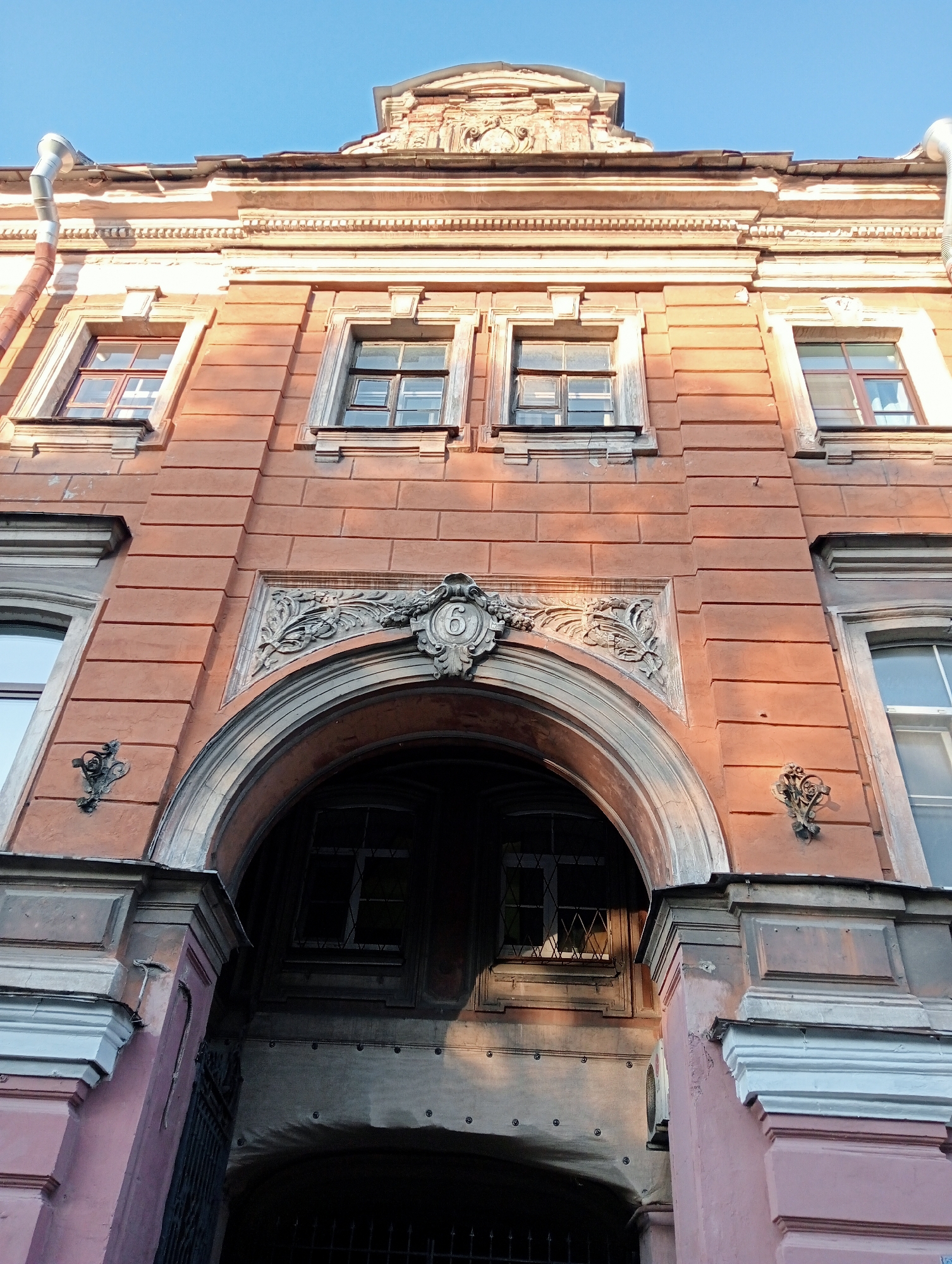 The image shows a low-angle view of the facade of an old, multi-story building, likely an apartment or commercial building, in Saint Petersburg, Russia. The building's exterior is painted a faded terracotta or salmon color with white architectural details.
Dominating the center of the image is a large, arched entryway. Above the arch, there is a decorative stone frieze featuring floral and scrolled motifs, and in the center of this frieze is a circular medallion with the number "6" carved into it. The arch itself is constructed of stone with visible layered stones and is supported by columns or pilasters that are partially obscured by shadow.
On either side of the arched entryway, and above it, are several windows. The windows are multi-paned and appear to be old, with some showing signs of wear and tear. The upper portion of the building features a more ornate cornice and decorative elements at the rooftop level, indicative of historical architecture. A metal downspout runs vertically along the upper left side of the building.
The lighting suggests it is daytime, with the sun casting shadows that highlight the architectural features and textures of the building. The sky above is a clear, pale blue. There are no people or animals visible in the frame. The overall impression is one of aged, classic European urban architecture.