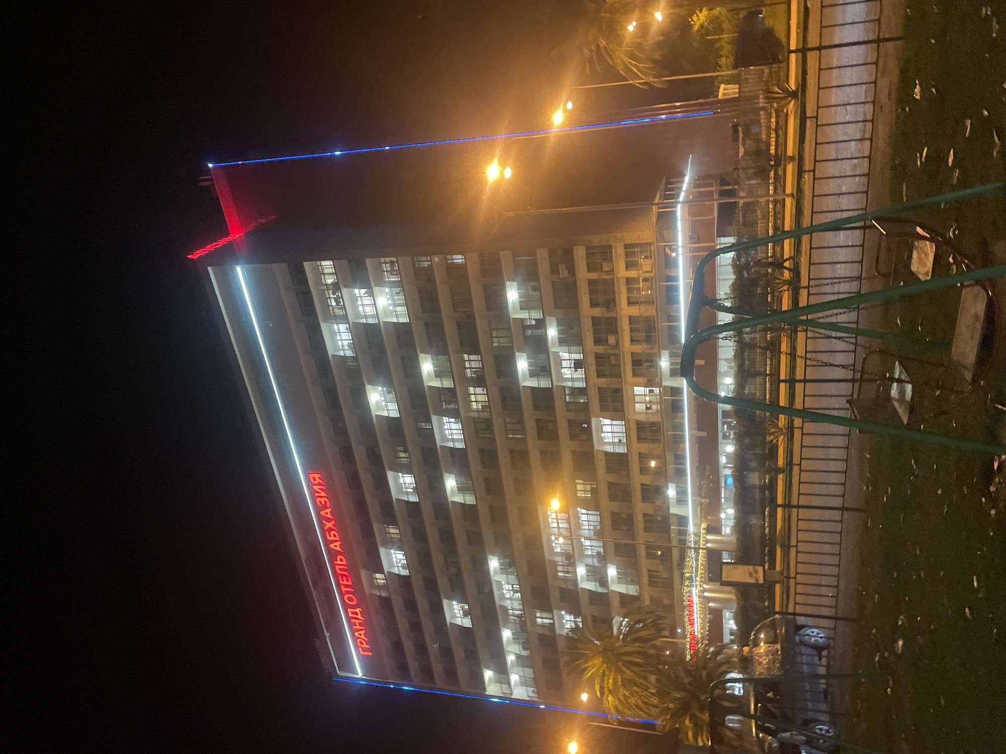 An outdoor nighttime shot shows a tall, multi-story hotel with illuminated windows and a neon sign. The sign, in red, reads "ГРАНД ОТЕЛЬ АБХАЗИЯ" which translates to "GRAND HOTEL ABKHAZIA". The hotel is located in the city of Gagra, Abkhazia. A red, angular neon structure is on the roof of the hotel, and a blue neon strip runs vertically down the side of the building. Several streetlights are visible, casting a warm glow. In the foreground, there is a small park with a swing set and some fallen leaves on the ground. A dark-colored car is parked on the street behind a metal fence. Palm trees are visible to the left and right of the hotel.