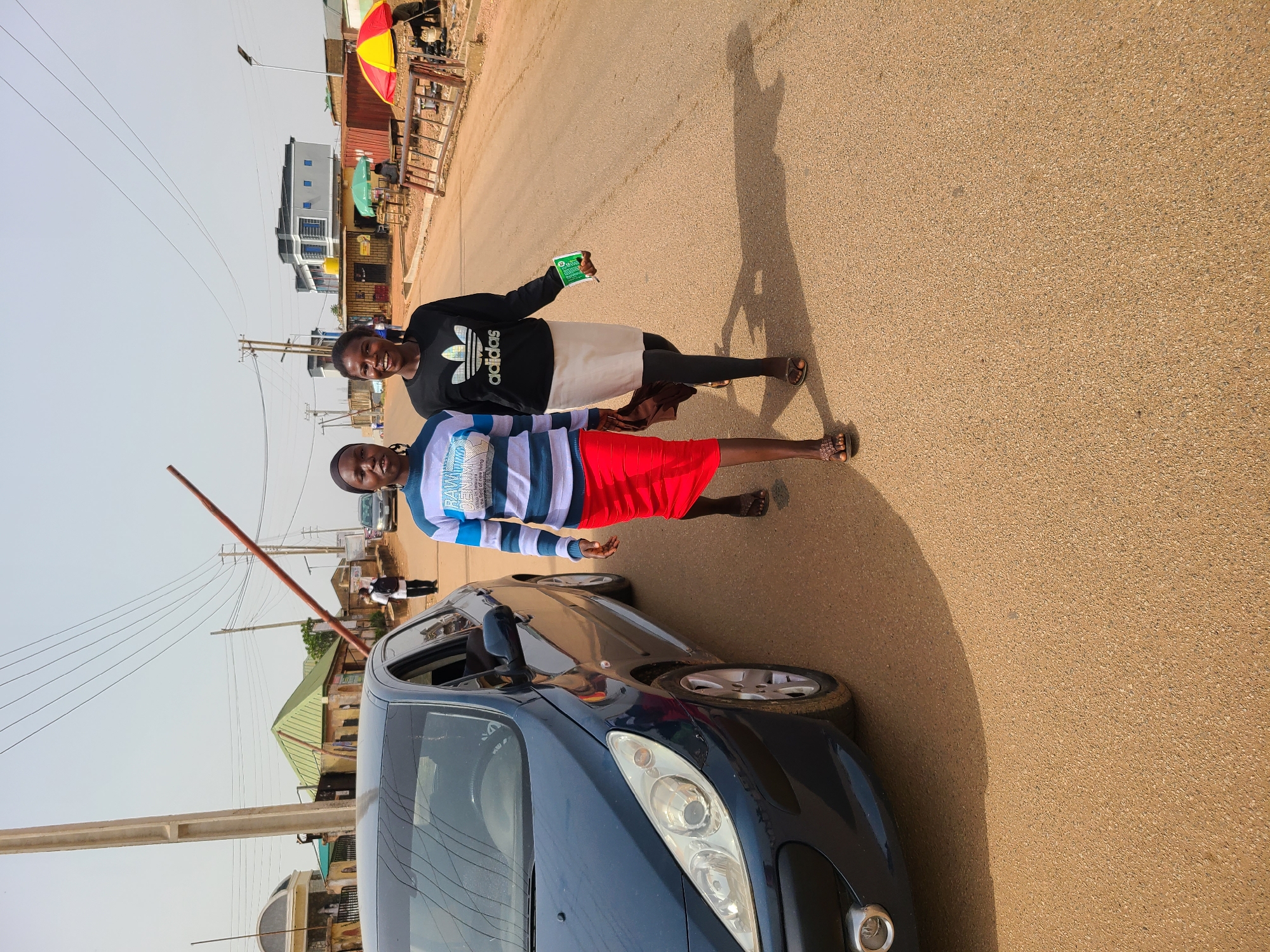 Two young women stand next to a dark blue car on a paved road in Jos, Nigeria. The woman closest to the camera wears a striped blue and white shirt and a red skirt. The other woman wears a black Adidas sweatshirt and a white skirt. Both women are smiling. The background includes buildings, shops, utility poles, and overhead wires, typical of an urban environment. A red and yellow umbrella-covered kiosk can be seen further down the road. The lighting suggests it is daytime. Shadows of the women are cast on the road.