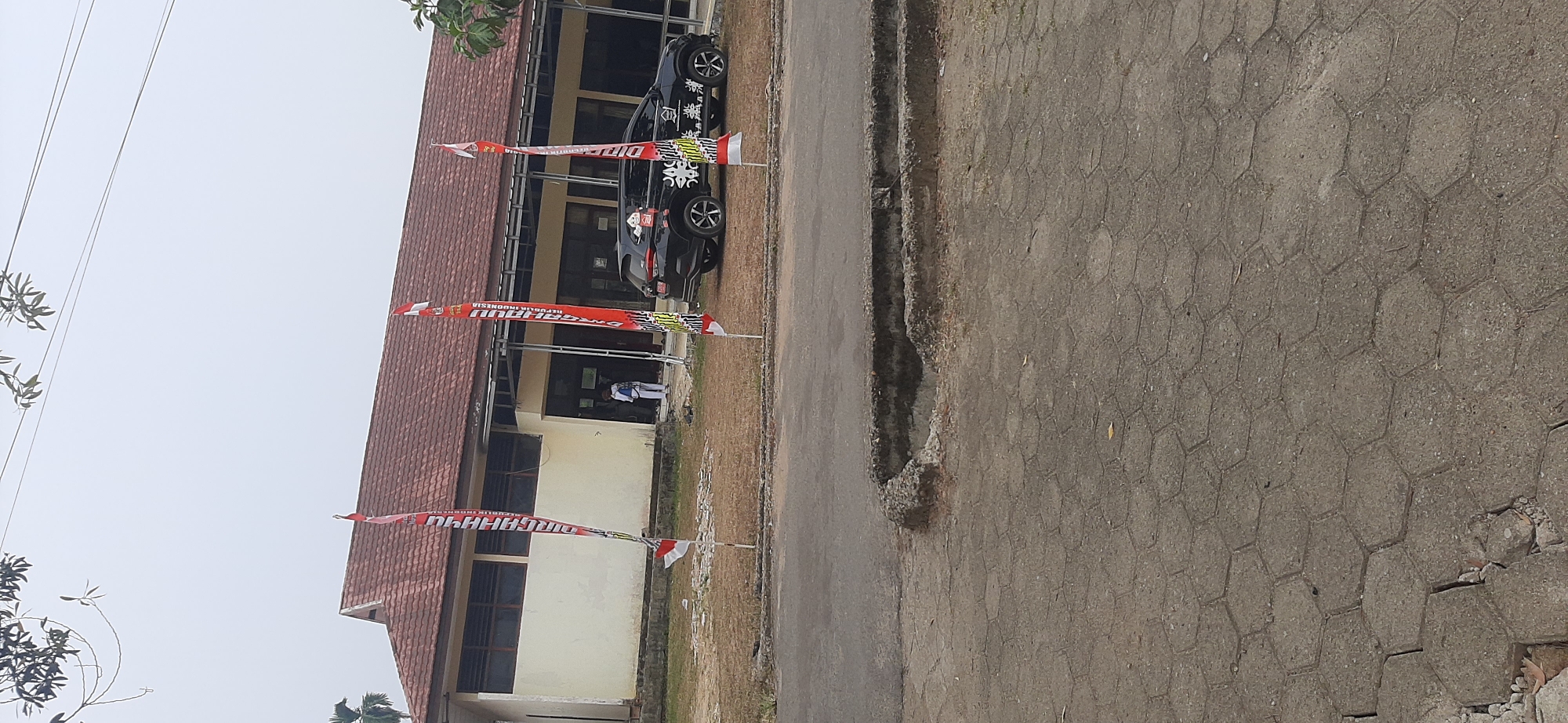The image depicts the exterior of a building, likely a school or government office, with a red tiled roof and cream-colored walls. In front of the building, several red and white flags are planted, some with text in Indonesian. A black car is parked on the right side of the building. One person, wearing white clothing, is visible standing in the doorway of the building. The foreground shows a paved area with hexagonal interlocking concrete blocks, and a section of road with a damaged concrete curb. Trees and power lines are visible in the background. The sky is overcast. This scene is consistent with the general appearance of public buildings and streetscapes found in Jambi City, Indonesia.