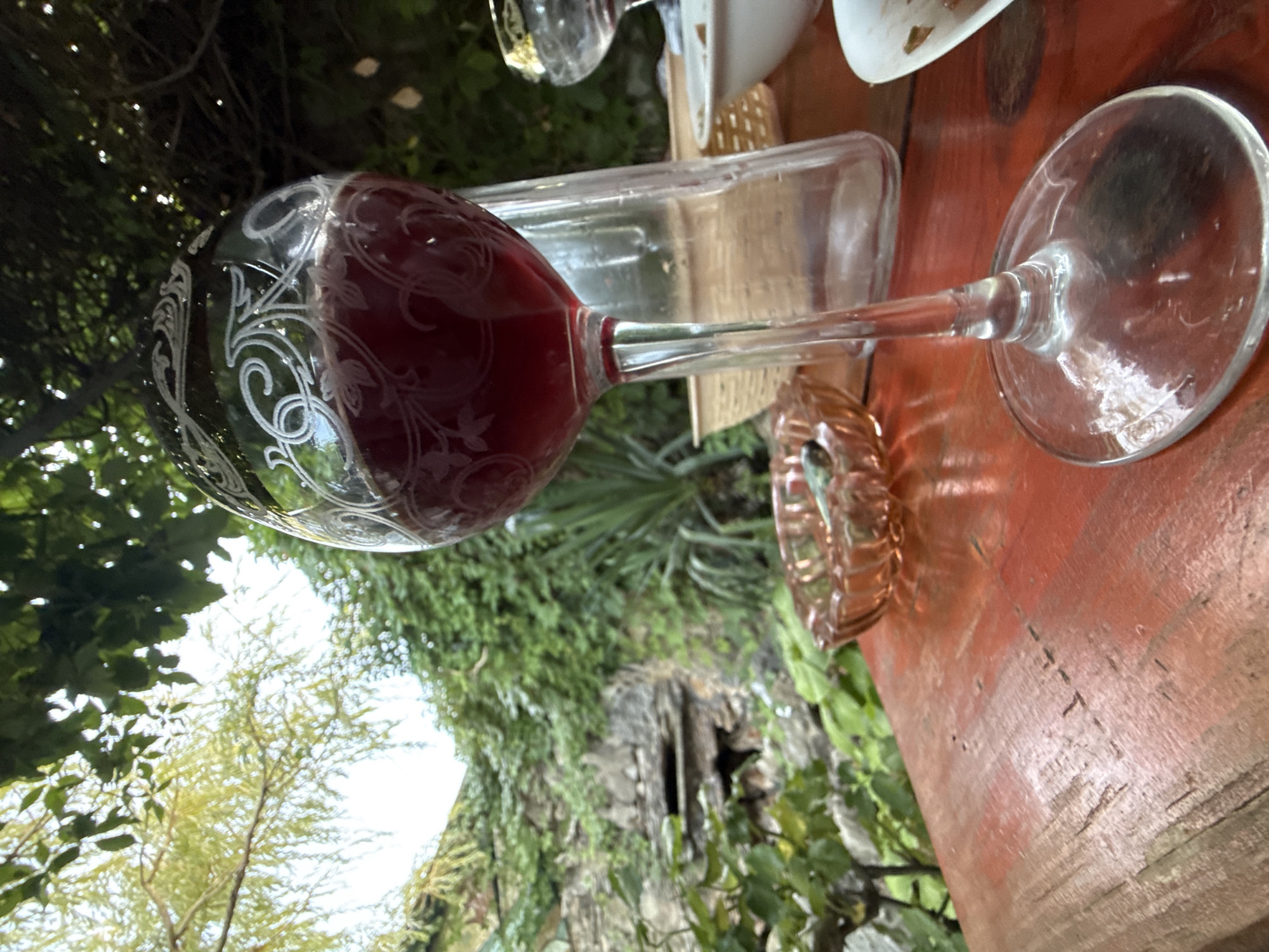 A close-up, low-angle shot captures a glass of red wine on a rustic wooden table. The wine glass, adorned with decorative white etchings, is filled with a deep red liquid. In the background, lush green foliage and an ivy-covered stone wall create a natural, almost overgrown atmosphere, suggesting an outdoor setting. A clear glass bottle, an ornate pink glass ashtray with cigarette butts, and another glass are also visible on the table. The scene implies a moment of relaxation or enjoyment, possibly in a garden or outdoor dining area. The location in Kerch, Ukraine, is not visually evident from the provided image.