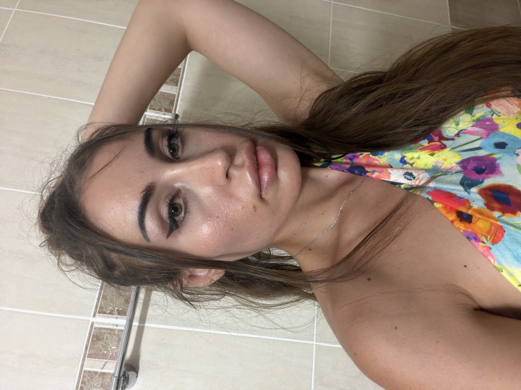 A medium shot captures a young woman with long, brown hair styled loosely, her left arm raised behind her head. She is facing the camera, her gaze directed forward. Her makeup includes winged eyeliner and lip gloss. She is wearing a colorful, floral-patterned garment. The background consists of tiled walls, suggesting an indoor setting. There is a towel rack visible on the wall. The image appears to have been taken in Tersefanou, Cyprus.