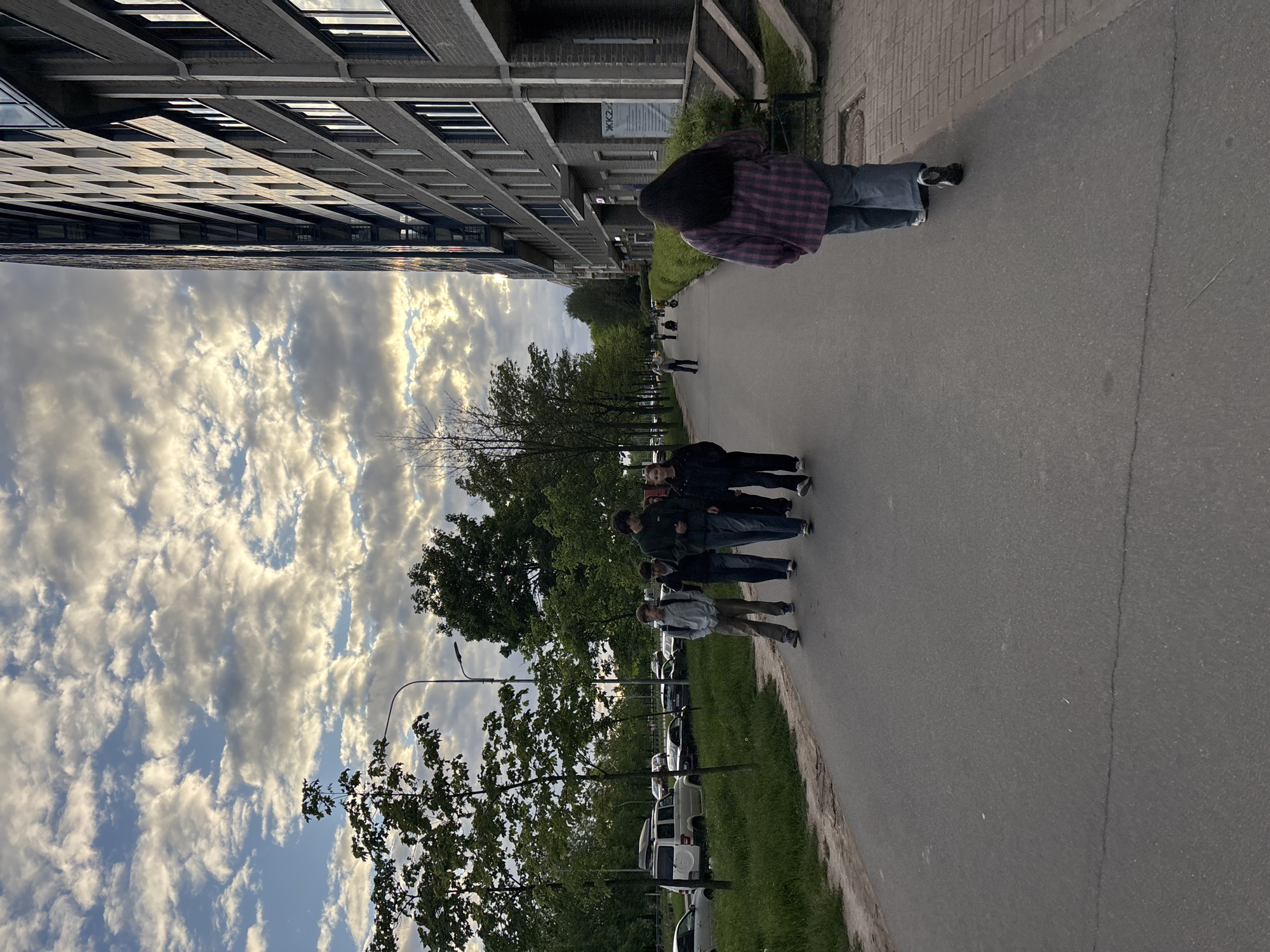 Here's a detailed description of the image:
**Setting:** The photo appears to be taken on a street or sidewalk in an urban area. On one side, there is a tall, multi-story building with a repeating pattern of windows. Across the street, there's a row of parked cars beside a grassy area and trees. The sky is partly cloudy with a mix of blue sky and white clouds. Streetlights are visible along the sidewalk.
**People:** There are several people walking on the sidewalk. One person is walking towards the camera wearing a plaid shirt and jeans. Further down the street, there is a group of approximately four people walking together. Another individual is further in the background walking away from the camera.
**Objects:** Parked cars are present on the right side of the image. Trees line the street. Streetlights are placed along the sidewalk. The building is a prominent feature of the scene.
**Actions:** People are walking on the sidewalk. Cars are parked.
**Interactions:** People are walking along the street in groups and individually.
**Overall Impression:** The image shows a typical street scene with a mix of residential/commercial buildings, trees, parked cars, and people. The lighting suggests it's daytime.