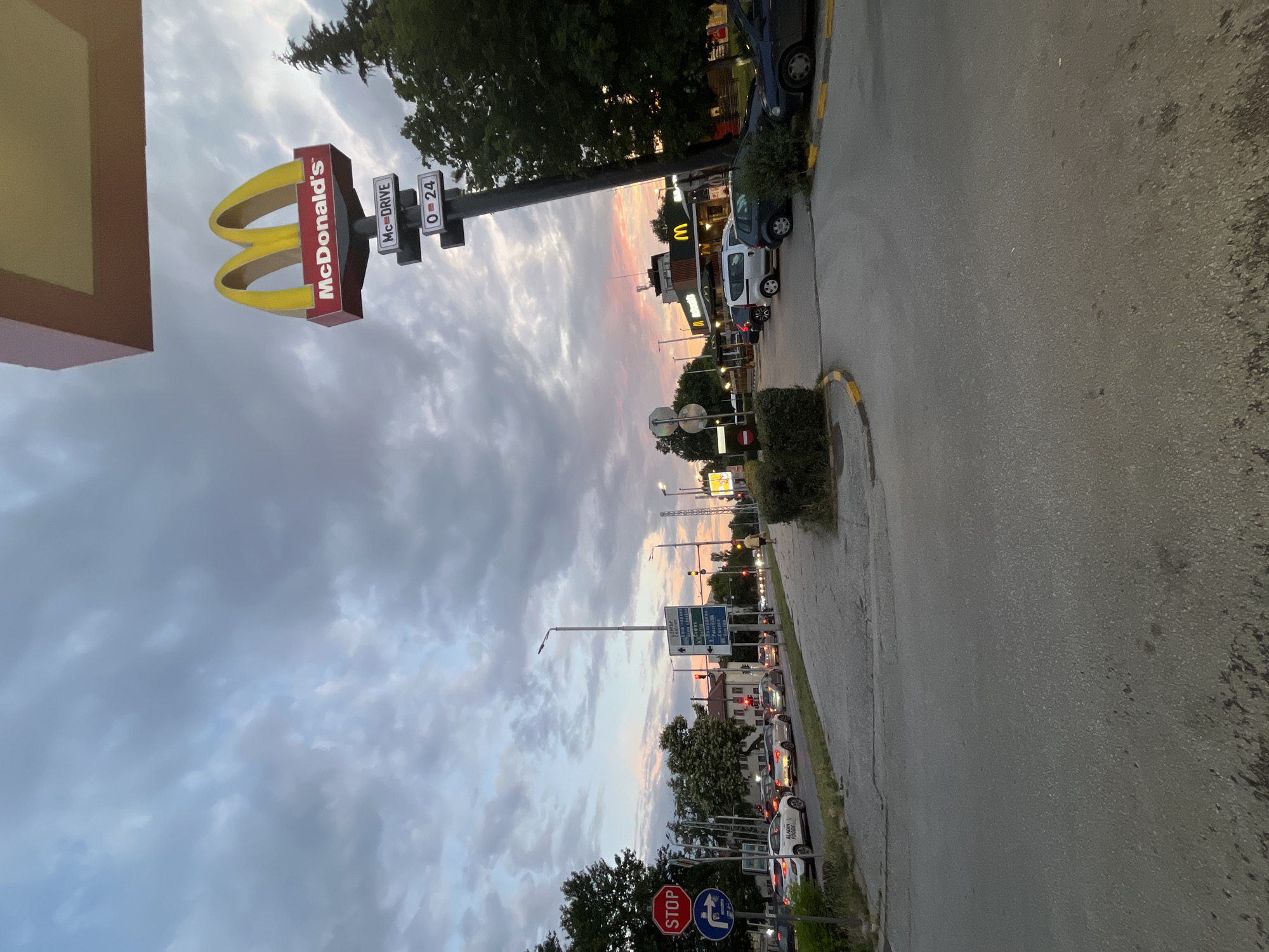 The image captures an exterior view of a McDonald's restaurant at dusk in Plovdiv, Bulgaria. A prominent McDonald's sign, featuring the iconic golden arches, is visible on a tall pole, with smaller signs indicating "McDRIVE" and operating hours "0-24". The sky is filled with dramatic, cloudy formations painted with hues of pink and orange from the setting sun. In the foreground, a paved road and sidewalk are present, with several cars parked and driving. The McDonald's building, with its distinctive architecture, is situated in the mid-ground. To the left, a busy intersection is depicted with traffic lights and road signs, including a red STOP sign. A person is walking on the sidewalk in the mid-ground, heading away from the camera. The scene is characterized by the interplay of artificial lights from the restaurant and streetlights against the natural light of the twilight sky.