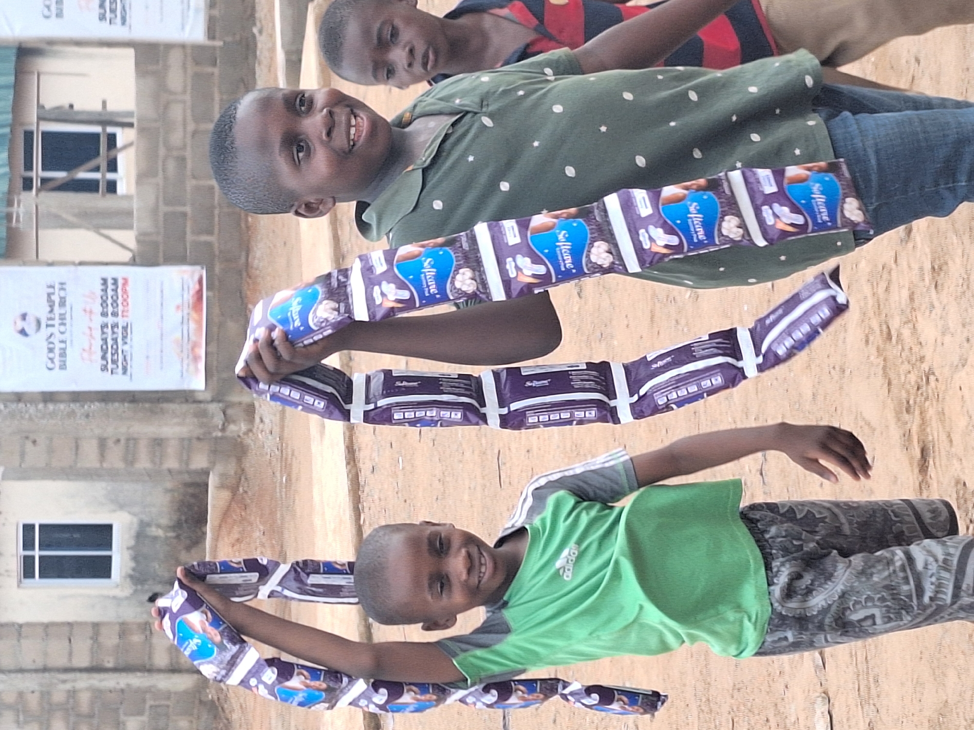 The image shows three children outside in Sabon Tasha, Nigeria. Two of the children are holding a chain of "Softcare" sanitary pad packages. One child is wearing a green shirt with the Adidas logo and grey patterned pants. Another child wears a polka dot green shirt and jeans. A third child wears a striped shirt. A building is in the background with a window and a banner for God's Temple Bible Church. The ground is dirt.