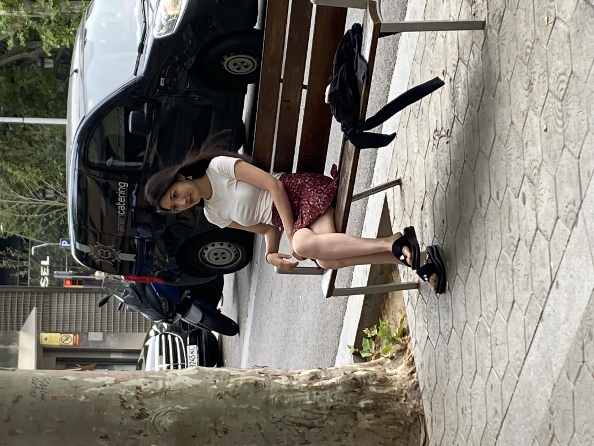 A young woman sits on a public bench on a street in Barcelona, Spain. She has long, dark hair, and is wearing a white t-shirt, a red floral print skirt, and black sandals. A black jacket is draped over the back of the bench. To the left, a black van with the word "arencatering" printed on the side is parked on the street. A motorcycle is parked behind the van. The base of a large tree is visible in the foreground. The license plate of the motorcycle shows "3245 KC". The street is paved with hexagonal tiles. The scene appears to be in daylight.