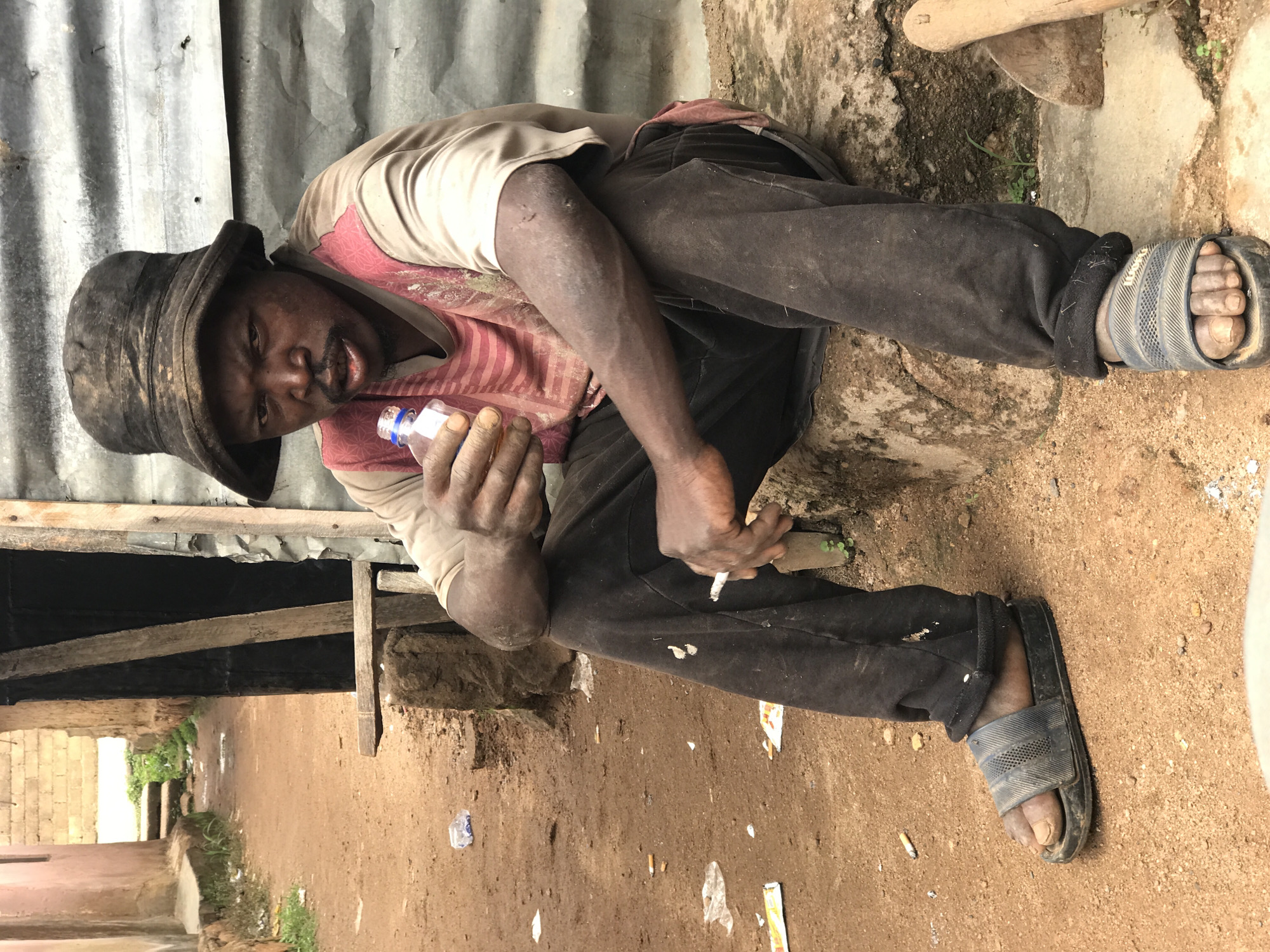 A man wearing a dirty, dark bucket hat and a t-shirt with red stripes and a beige collar sits on a concrete ledge. He holds a small clear bottle with a blue cap in his right hand and a lit cigarette in his left hand. He is wearing dark pants and dark blue slide sandals. The man is in an outdoor area with a dirt ground. In the background, there is a corrugated metal wall and a narrow path leading between buildings. Scattered on the ground are cigarette butts and some trash. The scene appears to be in an urban environment, likely a street or alleyway.