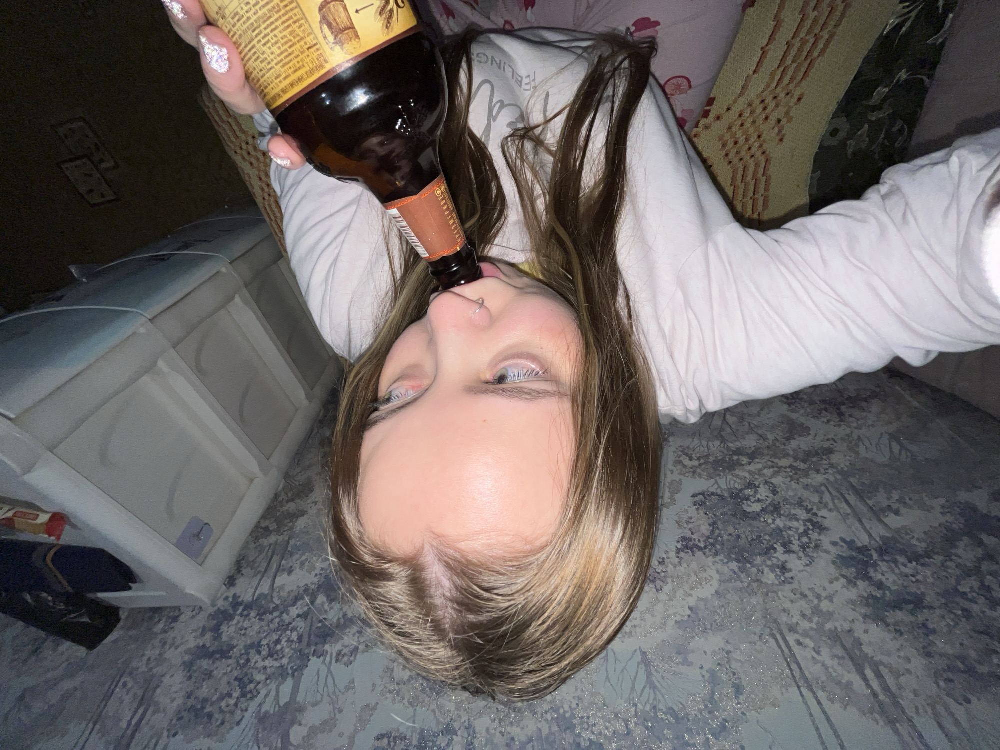 A young woman is lying on a bed, holding a dark brown bottle to her lips. She appears to be drinking from the bottle. She has fair skin, blonde hair, and blue eyes. Her nails are decorated with glitter. She is wearing a light-colored long-sleeved shirt. The bed has a floral patterned pillow and a gray and white patterned bedspread. To the left of the bed is a white plastic storage unit. The scene is poorly lit. There is no evidence of Novopokrovskaya, Russia.