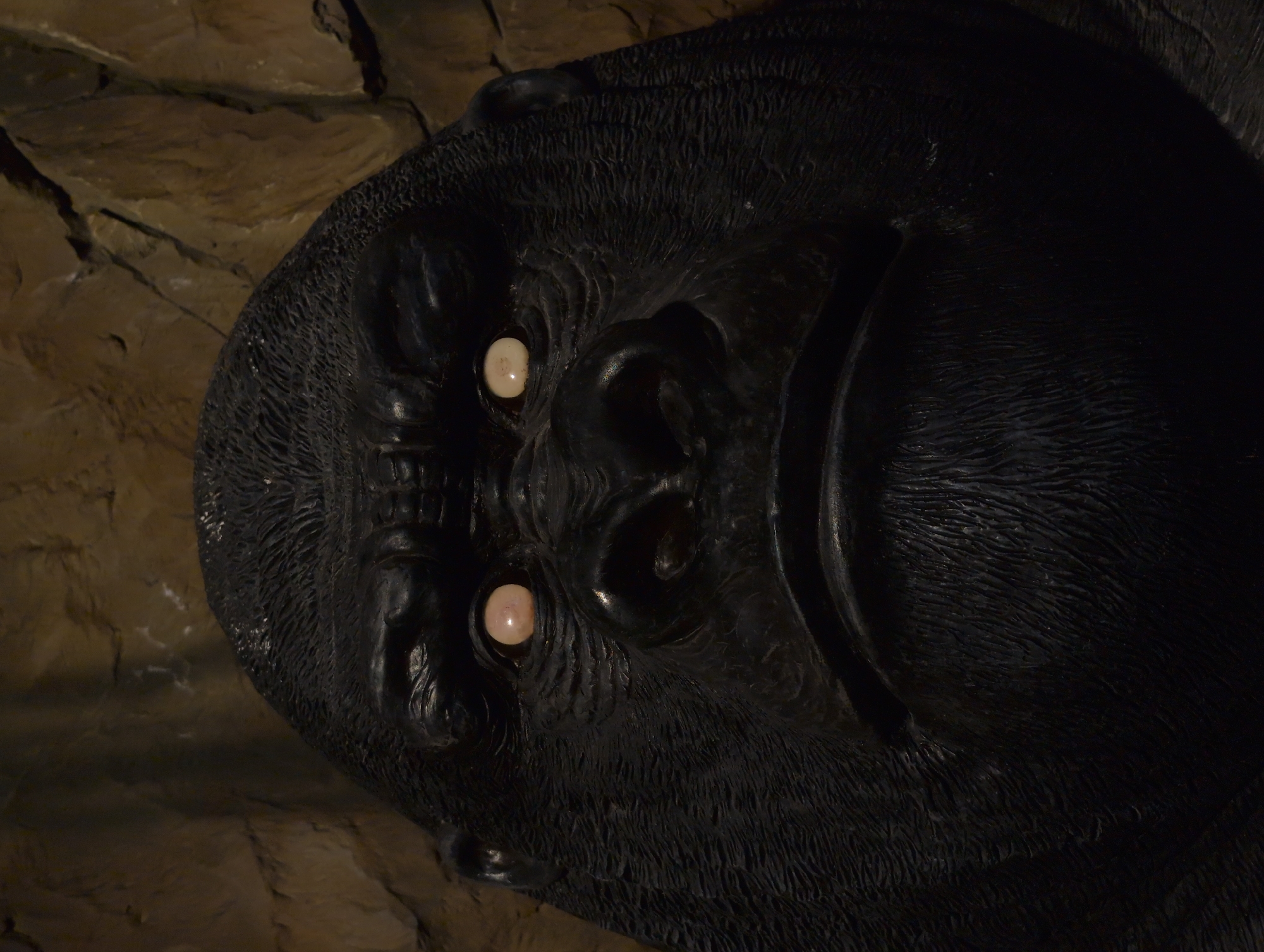 A close-up, low-angle frontal shot shows a dark, textured sculpture of a gorilla's head against a brown, rocky background. The gorilla's face is the primary focus, with its dark, leathery skin textured to resemble fur. Its eyes are pale, round, and slightly unsettling, giving the sculpture an eerie appearance. The gorilla's brow is furrowed, and its mouth is closed in a grimace. The rocky background, in muted earthy tones, appears to be a cave wall or similar natural formation. The lighting is dim, highlighting the contours of the gorilla's face and creating deep shadows. The image does not contain any information about the city of Gelendzhik, Russia.