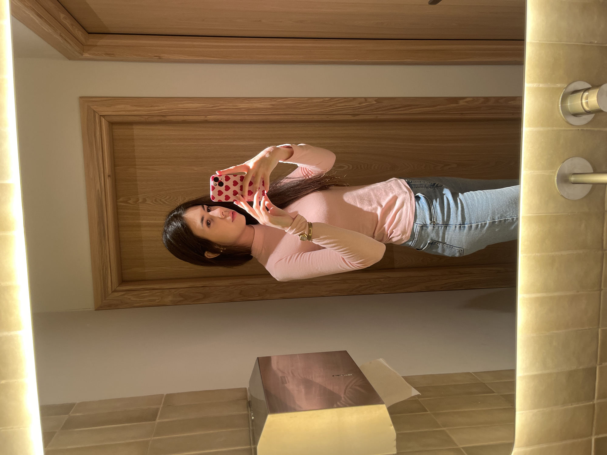 The image is a selfie taken in a bathroom with tiled walls. A woman is positioned in front of a mirror, holding a pink phone with a heart-patterned case. She is wearing a pink long-sleeved shirt and blue jeans. In the background, there is a wooden door and a tissue box on the bathroom counter. The mirror reflects the environment around the woman. The location is Unknown, Unknown.