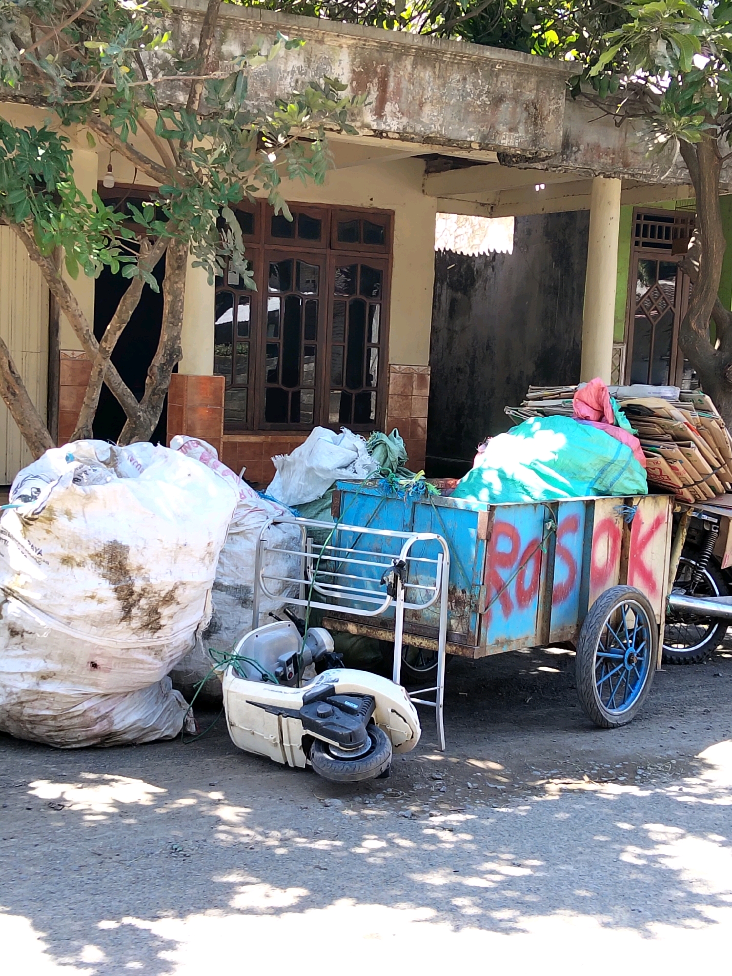 A two-wheeled cart filled with recyclable materials, such as plastic bags and cardboard, is parked on a dirt road in front of a weathered building. A white scooter lies on its side to the left of the cart. The building, likely a home, has brown wooden doors and windows, and is partially covered by trees with green foliage. The cart is painted blue with red lettering on its side that reads "ROSOK". The scene appears to be in Banyakan, Indonesia, based on the provided context.
