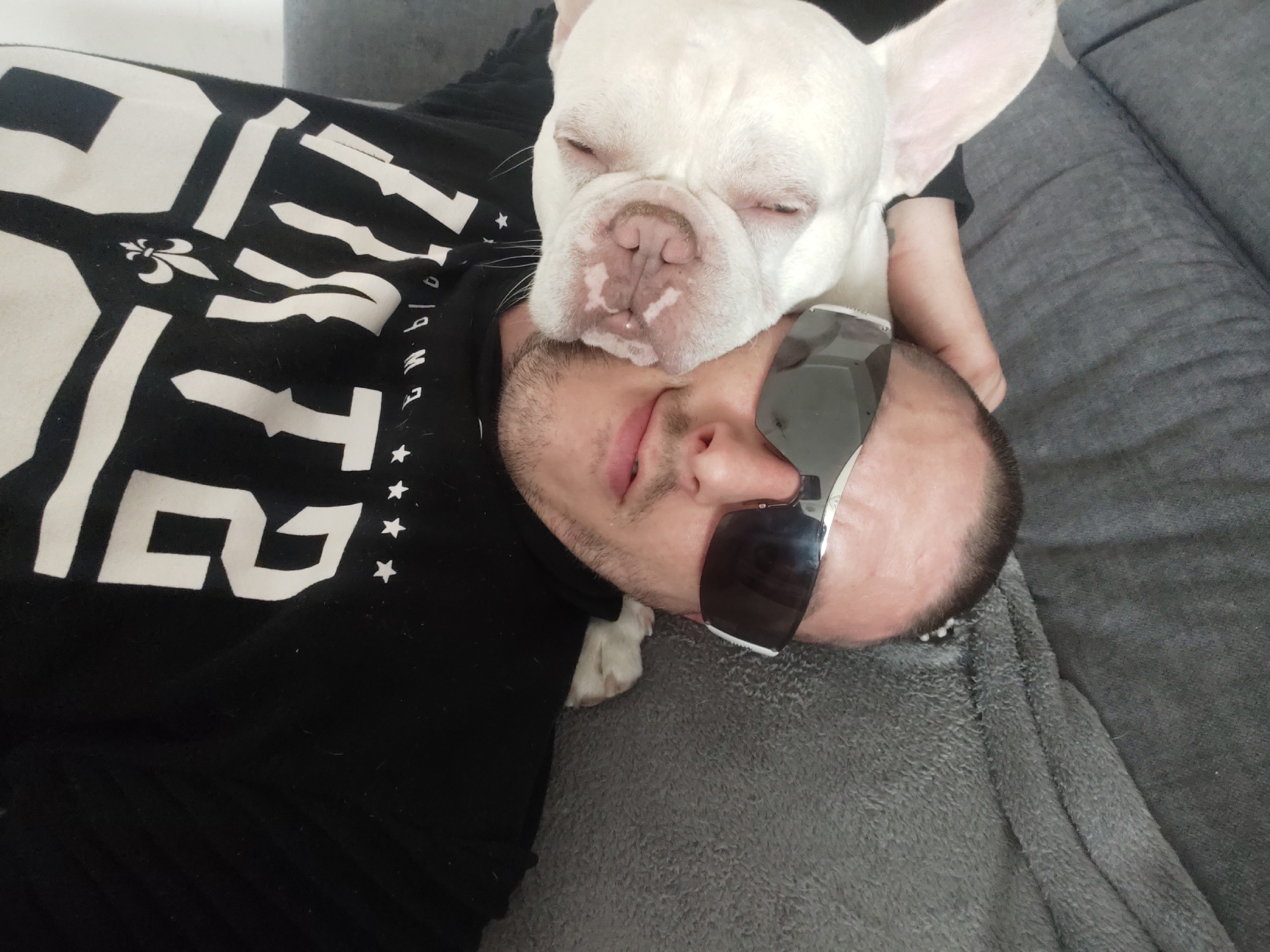 A close-up shot from an overhead perspective shows a man and a dog lying on a gray couch. The man, seen from the chin up, is wearing dark sunglasses and a black shirt with white designs on it. He has short, dark hair and a beard. A white French bulldog rests on the man's chest, with its head near the man's face. The dog's eyes are closed. Part of the couch is visible in the background. The location is not discernible from the visual content of the image.