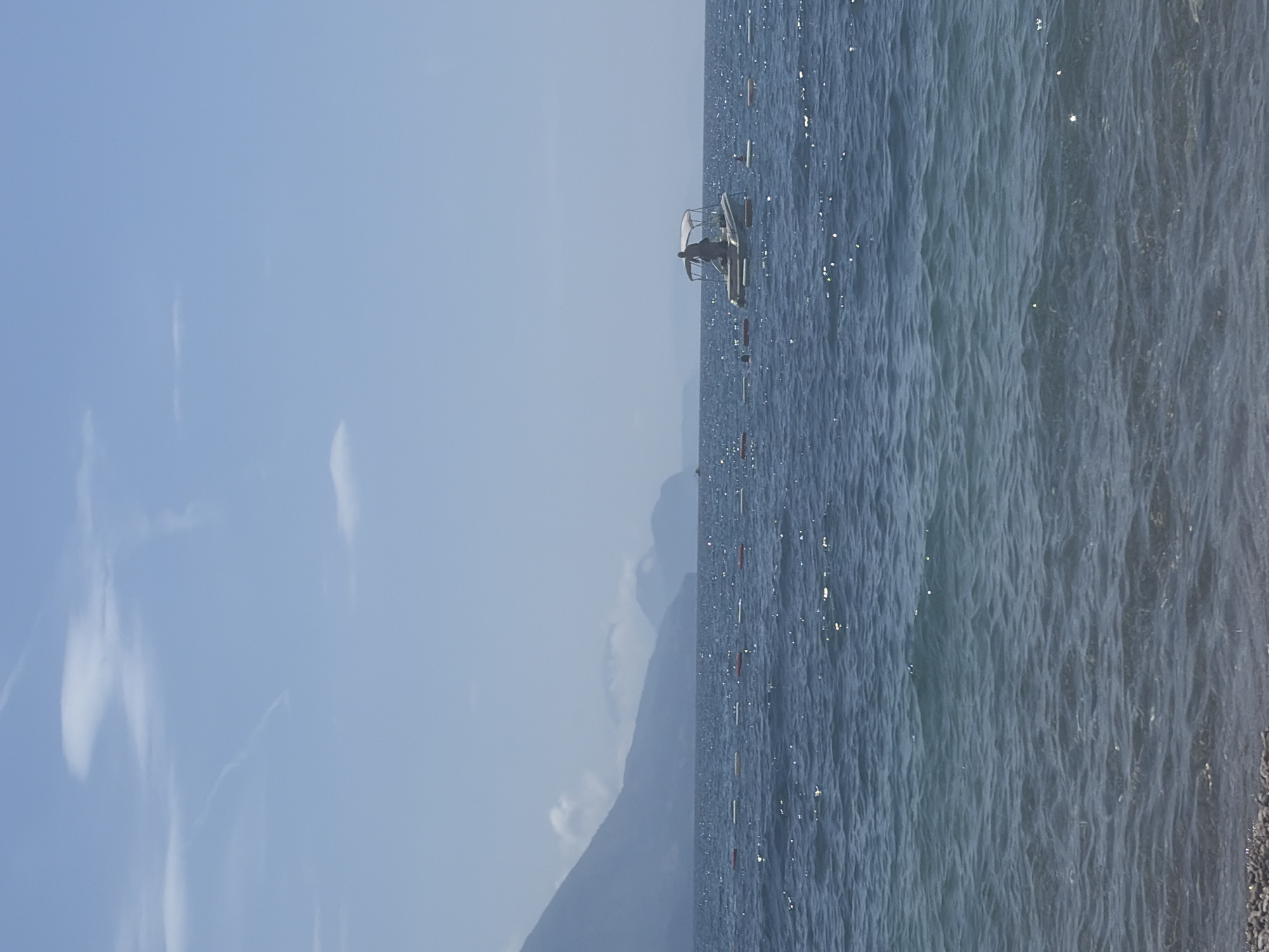 The image shows a boat on the water, with a person visible in the boat. The sky is a light blue with some clouds. In the background, a mountain is visible. The body of water appears to be a sea, with reflections on the surface. The location is not specified as being in Prijevor, Montenegro, and there is no visible indication of the city. Other objects include possible mooring buoys in a line.