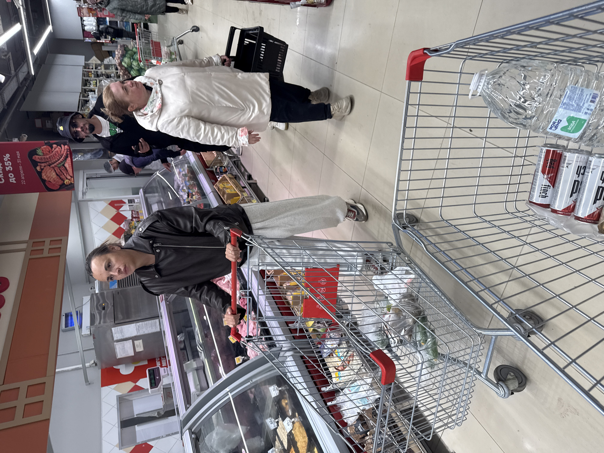 This image captures an indoor scene within a supermarket or grocery store in Yekaterinburg, Russia. Several people are engaged in shopping activities amidst aisles stocked with various products.
In the foreground, a person with dark hair tied back, wearing a black leather jacket and light grey sweatpants, pushes a metal shopping cart filled with groceries. The cart contains items in plastic bags and a red box. This person looks towards the camera with a neutral expression.
To the right, another metal shopping cart is partially visible, holding a large clear plastic bottle of water and several cans of what appears to be "Finch" brand beer. The water bottle's label clearly shows "МОЯ ЦЕНА" (MY PRICE) and "ВОДА" (WATER).
Further into the aisle, a woman in a white puffer jacket and dark trousers carries a black plastic shopping basket. She has blonde hair and looks upwards. Behind her, a man wearing a dark hat, sunglasses, and a black t-shirt stands near her. Other shoppers are vaguely visible in the background, interacting with merchandise or pushing carts.
The store features refrigerated display cases, visible on the left, containing various food items such as meat and sausages. Overhead, fluorescent lights illuminate the space, and the floor is tiled in a light color.
A prominent red promotional sign on the left advertises "СКИДКА до 35%" (DISCOUNT up to 35%) and specifies the dates "22 апреля – 31 мая" (April 22 – May 31), alongside images of sausages and grilled meat, indicating a sale on these products. The overall atmosphere is that of a typical, busy grocery shopping environment.