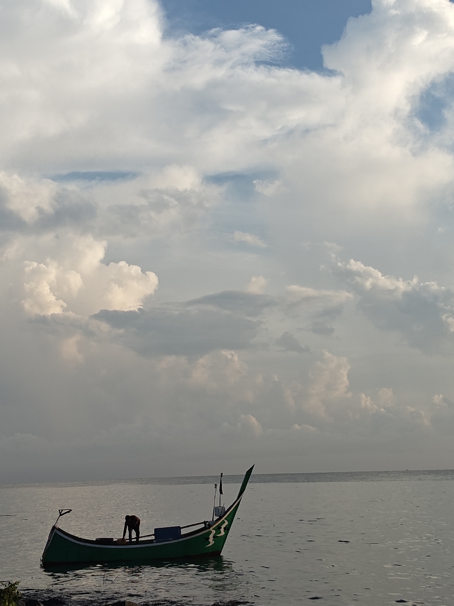 The image shows a green fishing boat in calm water, close to the shore. A single person is in the boat, appearing to be working or preparing something. The sky is filled with heavy, layered clouds, with patches of blue visible. The scene appears to be taken at the seashore in the city of Lhokseumawe, Indonesia.