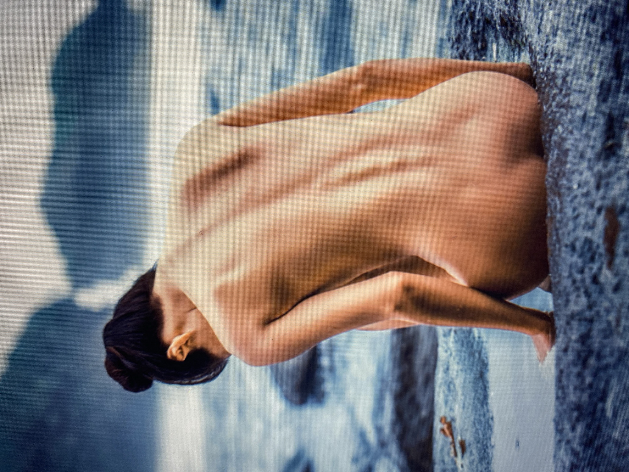 A person is captured from behind, kneeling on rocks by the water. Their back is prominently displayed, showcasing the contours of their spine and shoulders. The background is a blurry expanse of water and distant landforms, possibly mountains, shrouded in mist or haze, creating a soft, atmospheric setting. The overall impression is one of serene contemplation or connection with nature.