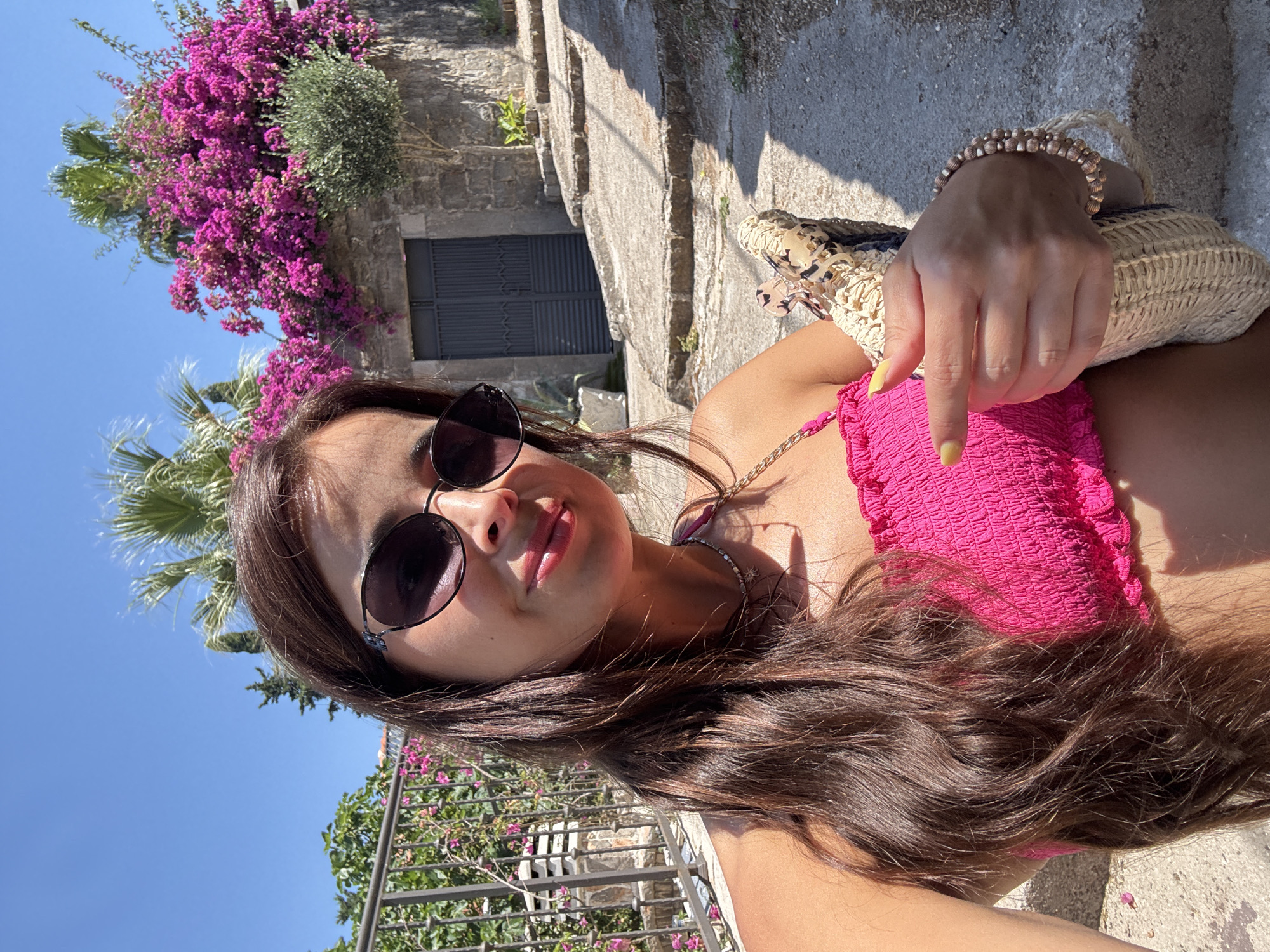 A young woman with long brown hair, wearing sunglasses and a pink smocked tube top, poses for a selfie outdoors. She is holding a woven bag in her left hand. Behind her, a stone building with a gray door is visible, surrounded by vibrant pink bougainvillea flowers and palm trees. The ground appears to be a sun-drenched stone pathway or stairs. The sky is a clear blue.