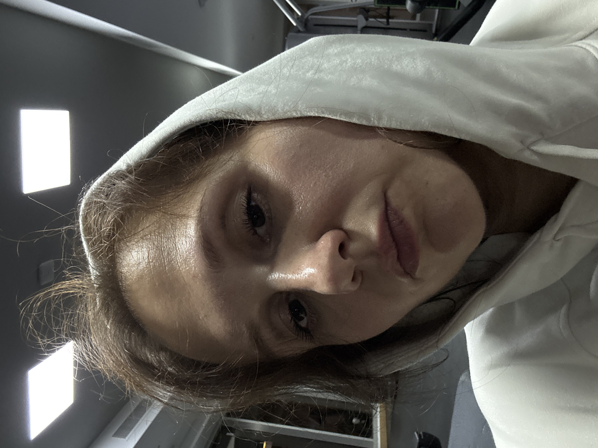 A close-up, slightly angled portrait shows a person with fair skin wearing a white hooded sweatshirt. The hood is up, framing their face. They have brown hair, visible around the edges of the hood, and dark eyes. Their skin appears slightly damp. The background is blurred, showing a grey wall with two rectangular light fixtures and some metallic structures overhead. No specific location in Dubai is discernable from the image.