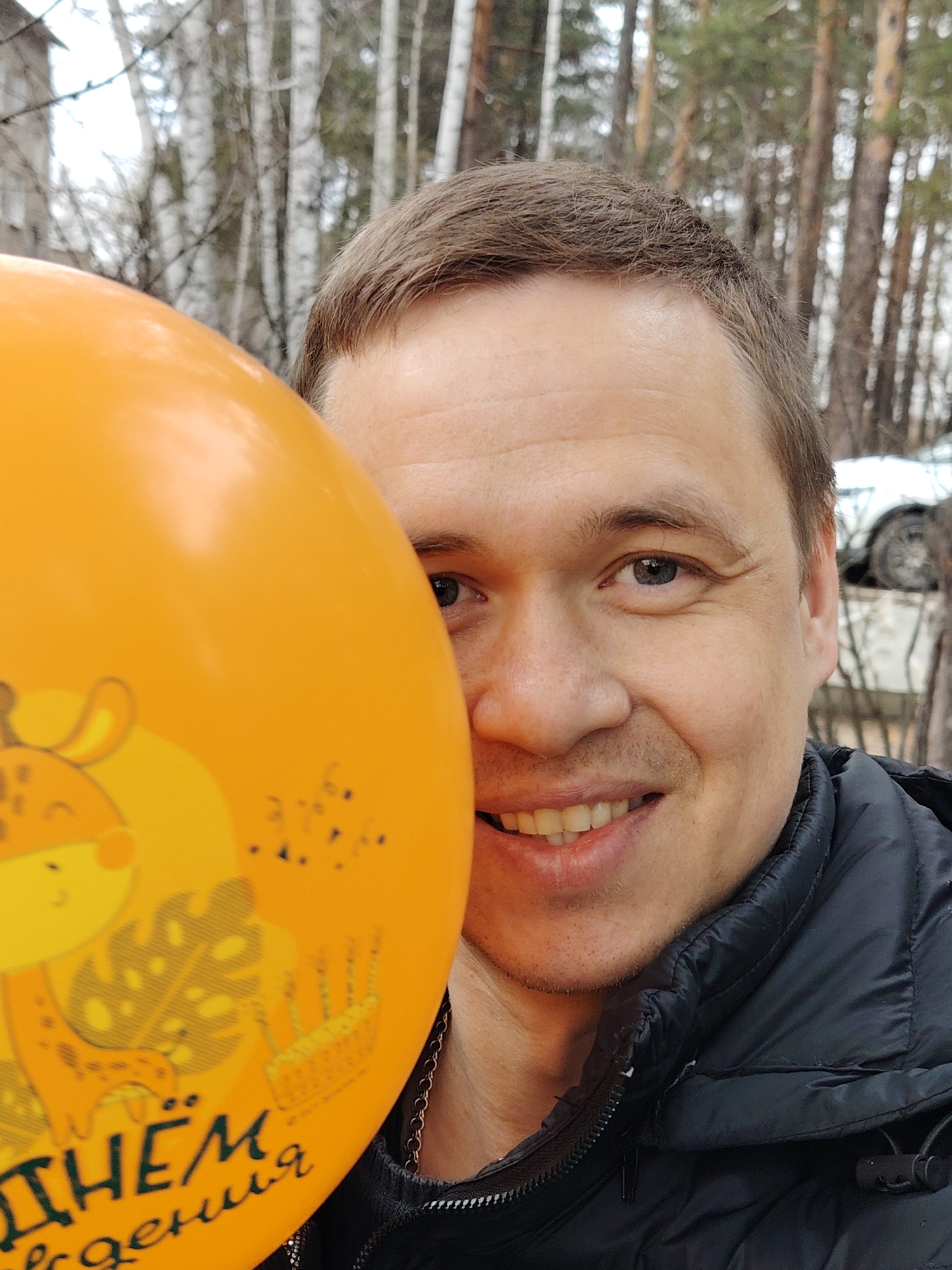 A close-up outdoor portrait captures a smiling adult man, partially obscured by a large orange balloon. The man has short, light brown hair, fair skin, and blue-gray eyes. He is seen with a wide, happy smile, showing his teeth, and faint smile lines are visible around his eyes. He is wearing a black puffer jacket with a zippered collar, and a metallic chain can be seen around his neck.
The prominent orange balloon occupies the left side of the frame, covering part of the man's face. It features a cheerful cartoon design of a giraffe wearing a party hat, surrounded by decorative leaves and a small birthday cake. Visible Cyrillic text on the balloon reads "ДНЁМ" (Day) followed by "рождения" (of birth), translating to "Happy Birthday."
The background indicates an outdoor setting, likely in Chaikovsky, Russia. A dense cluster of tall, slender trees, including distinctive white-barked birch trees and some conifers, forms a natural backdrop. The bare branches of the deciduous trees suggest a late autumn, winter, or early spring season. A light-colored building is partially visible on the far left, and a white car can be seen in the distant background on the right. The lighting is natural and diffuse, suggesting a daytime scene with overcast skies. The overall mood conveyed by the man's expression and the festive balloon is one of joy and celebration.